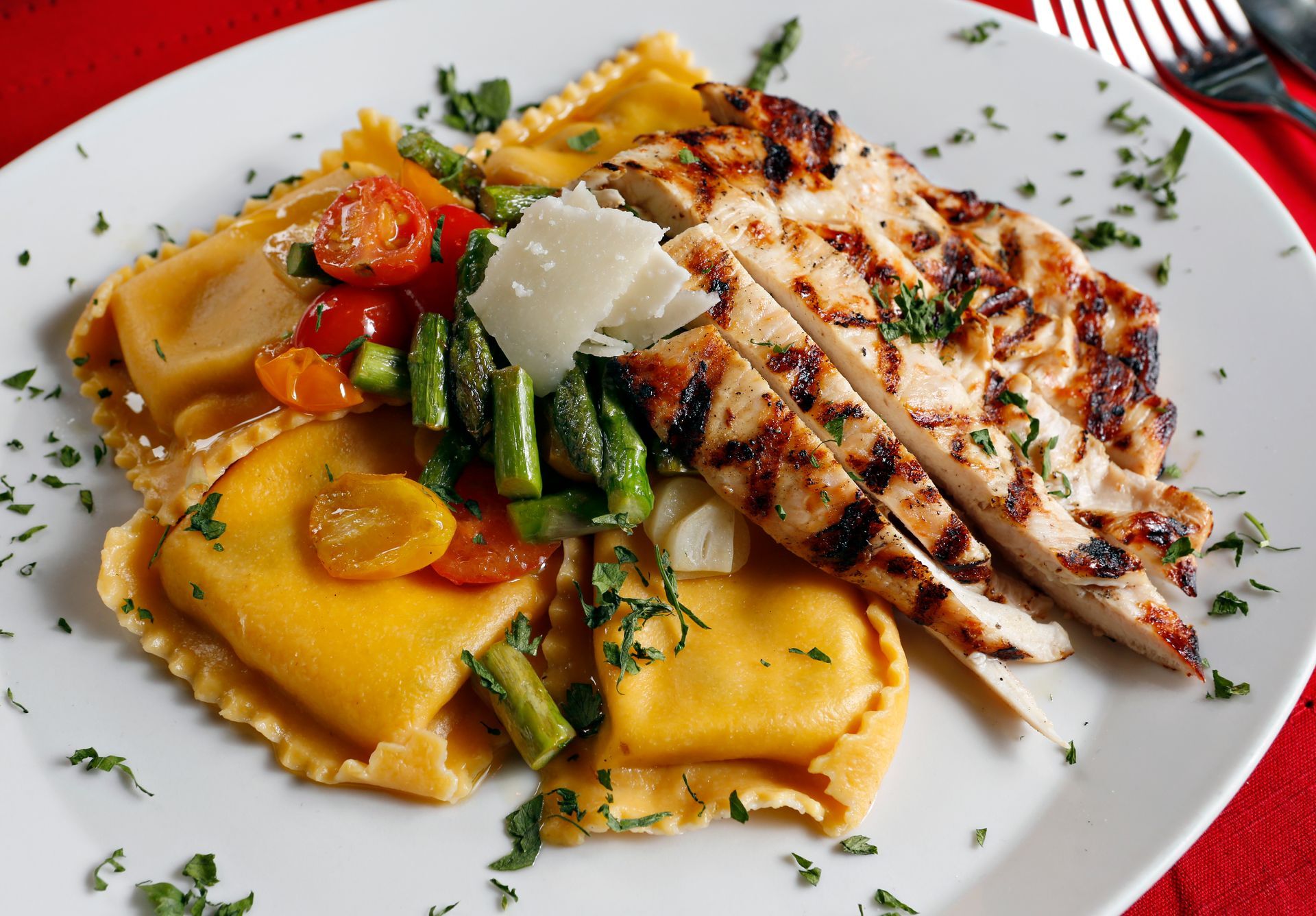 Grilled sliced chicken breast served with ravioli, asparagus, and cherry tomatoes on a white plate with herbs.