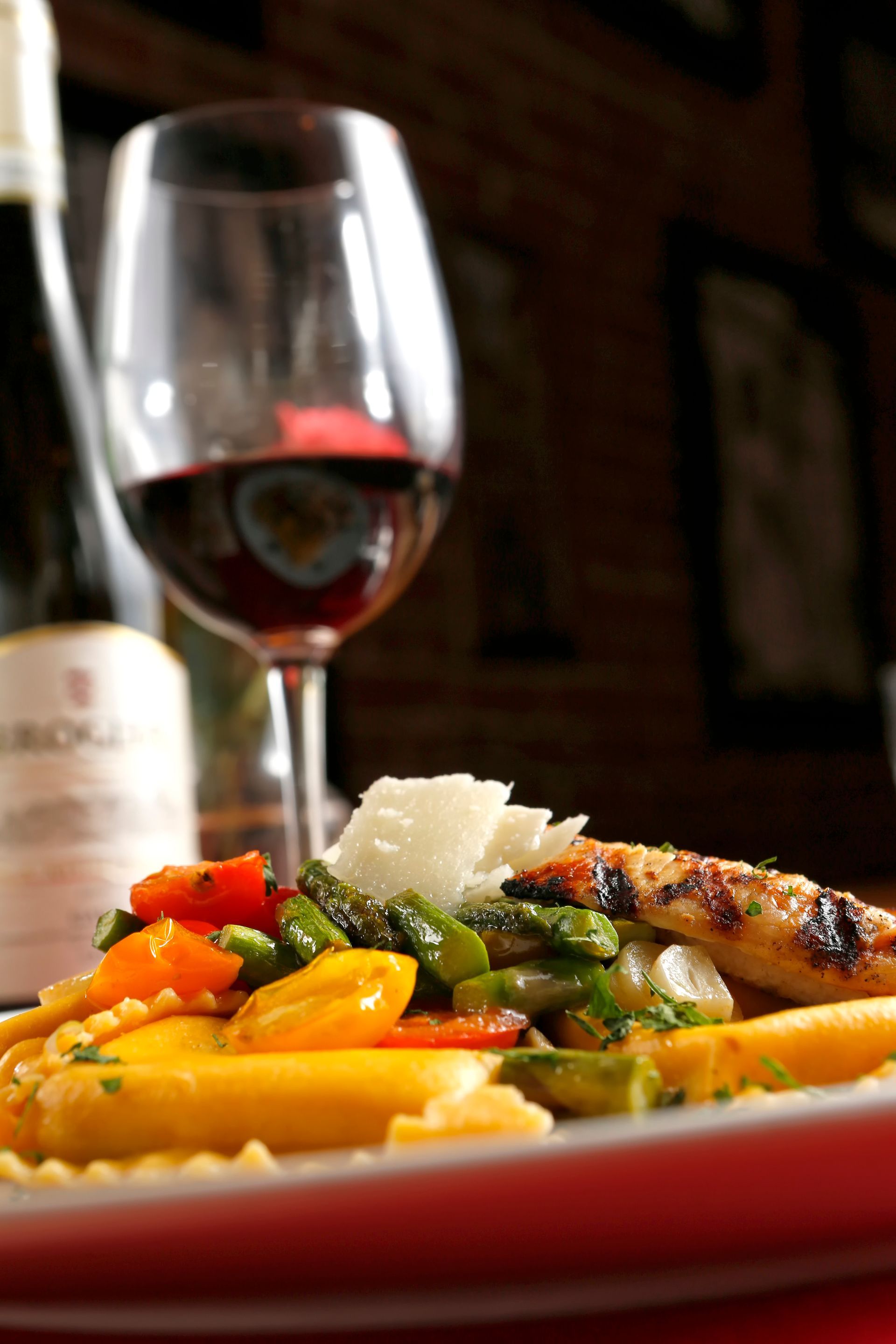 A plate of pasta with grilled chicken, asparagus, and tomatoes, next to a glass of red wine in a dim restaurant.