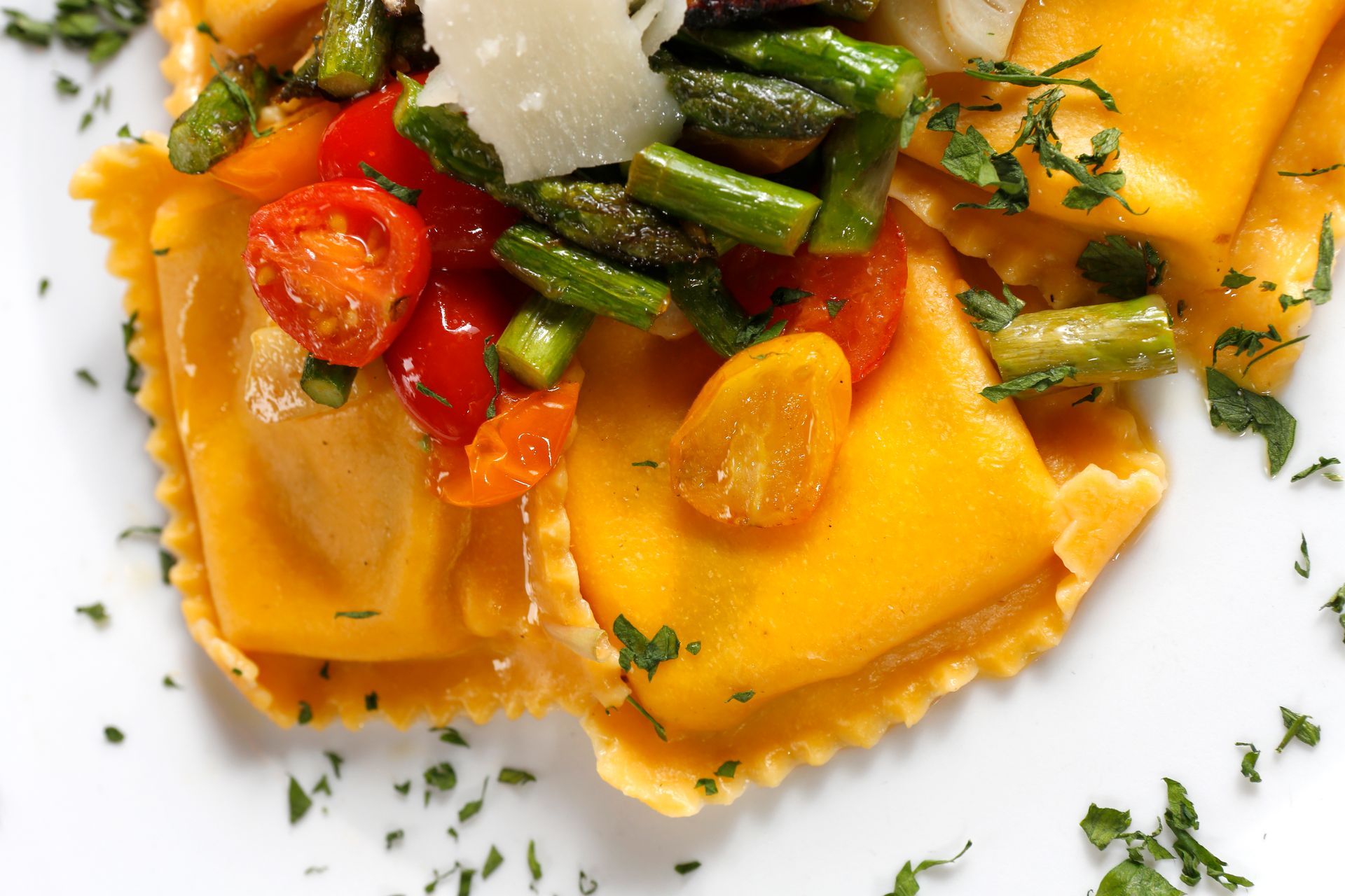Two ravioli squares topped with sautéed asparagus, cherry tomatoes, and shaved parmesan on a white plate.