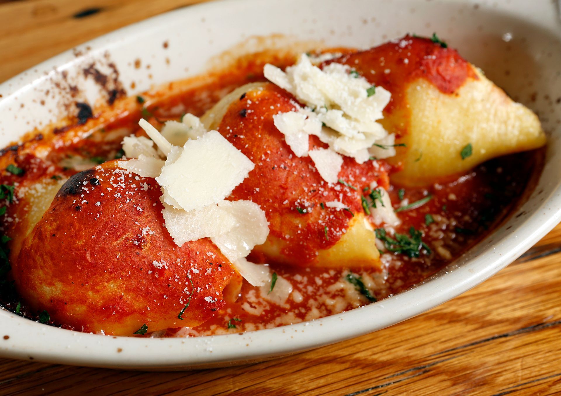 Three stuffed pasta shells topped with shaved parmesan and red tomato sauce in a white dish on a wooden surface.