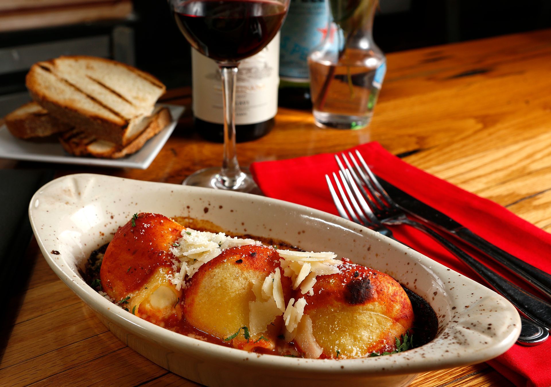 A dish of baked, saucy pasta topped with cheese in a white bowl, served with toasted bread and a glass of red wine.