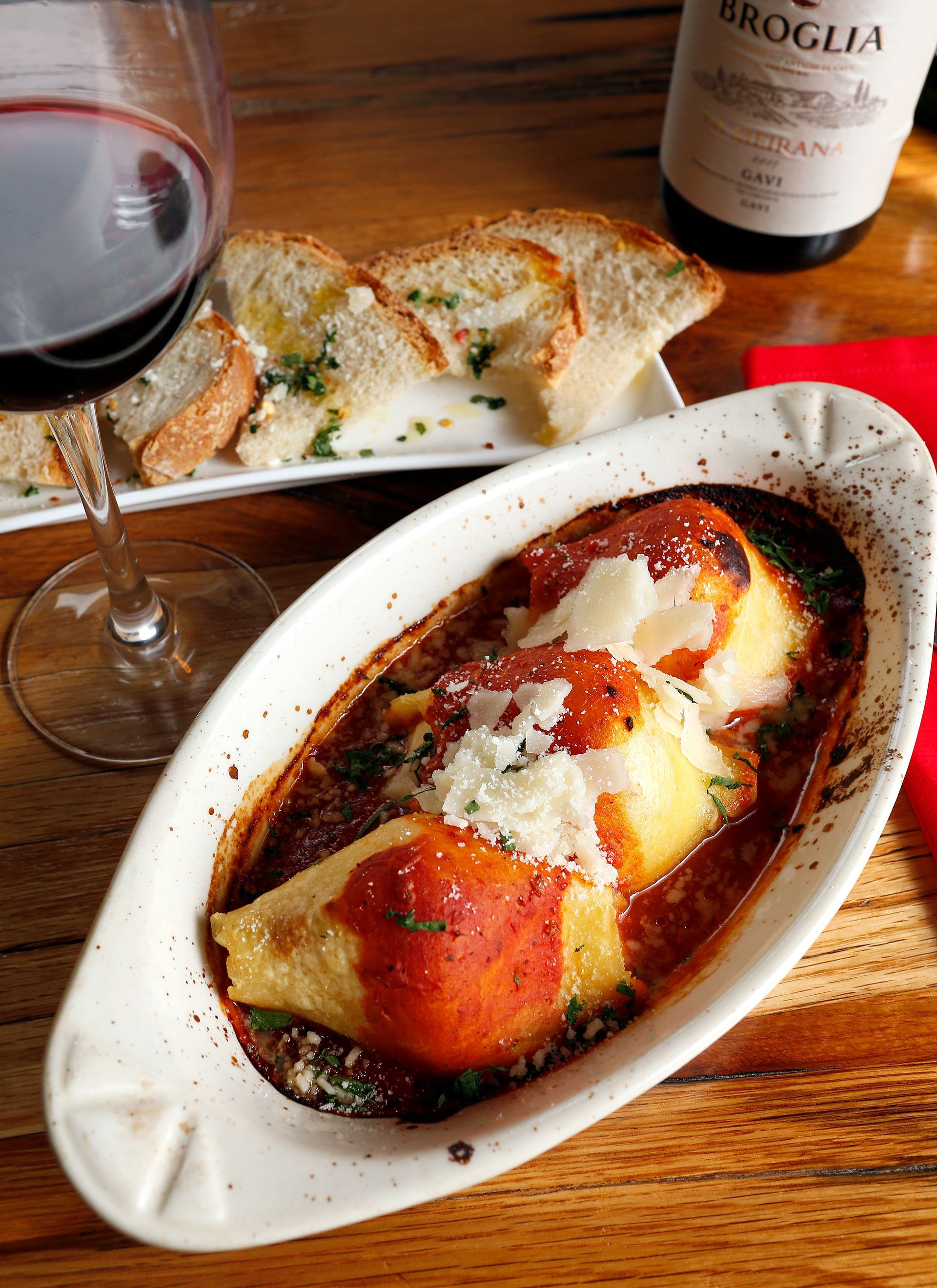 Baked stuffed pasta shells in tomato sauce, served with bread, a glass of red wine, and a bottle of wine on a table.