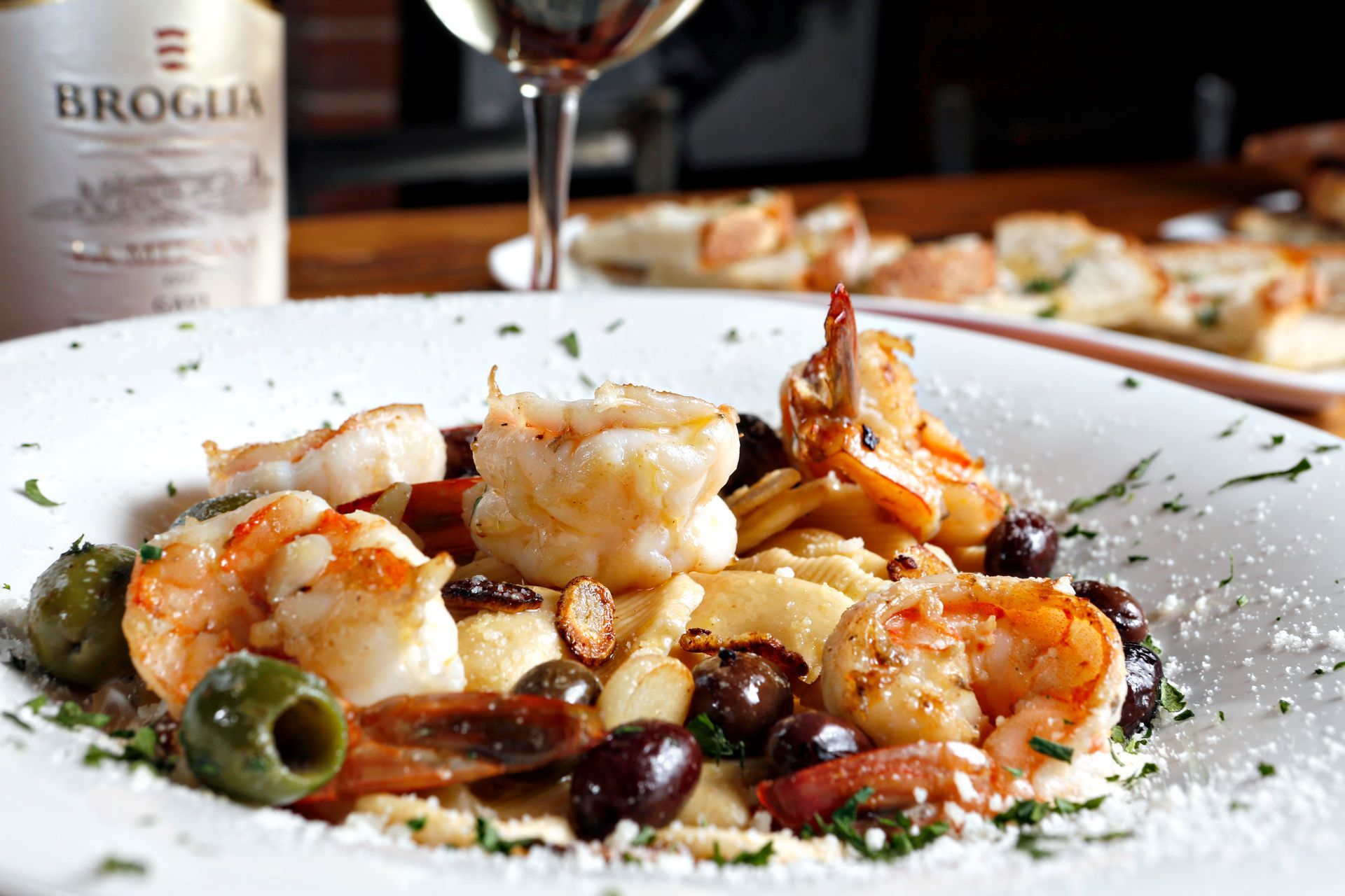 A white bowl of shrimp pasta with olives, topped with herbs and cheese, accompanied by a glass of wine and bread.