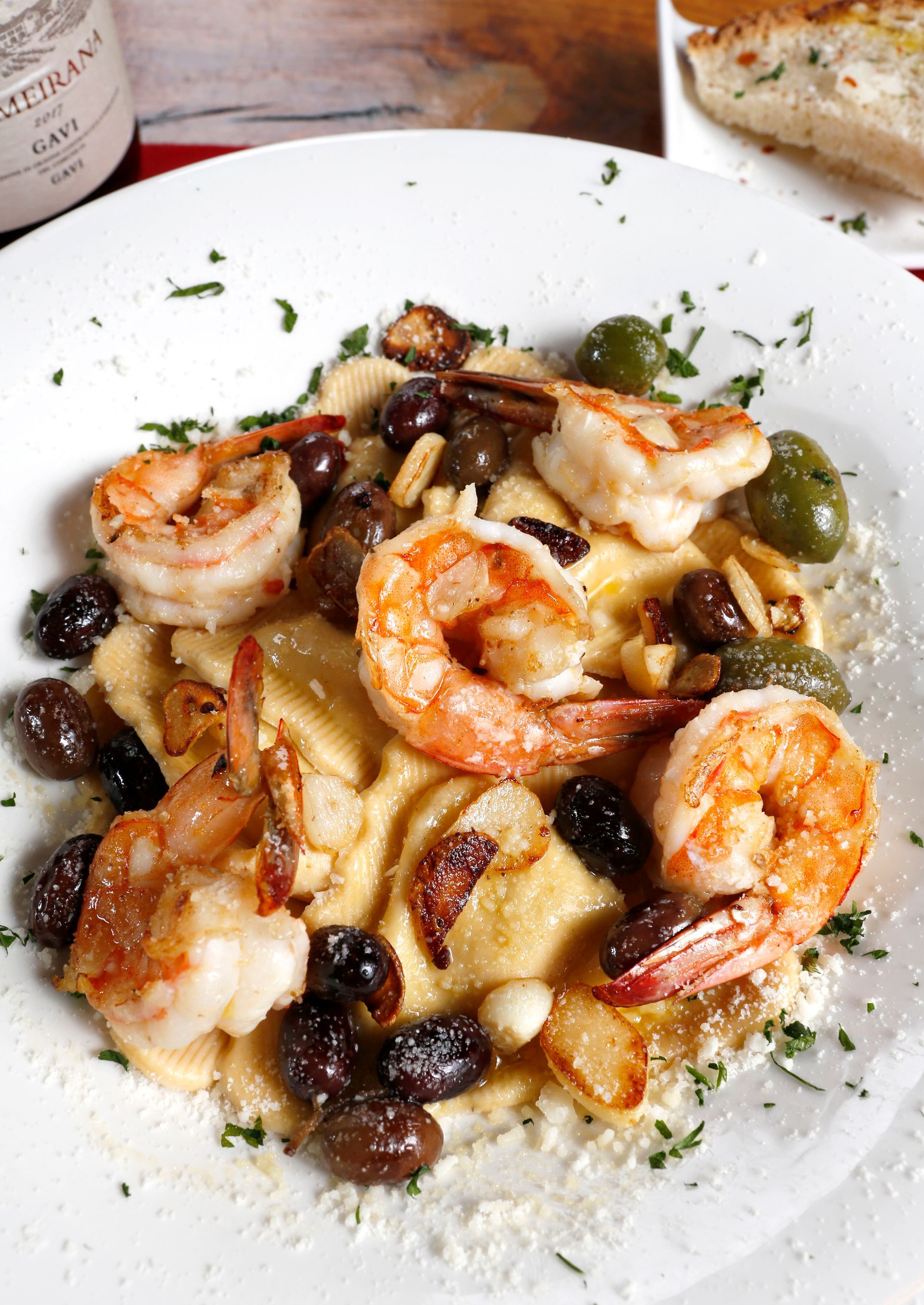 A white plate of seared shrimp over creamy polenta, garnished with olives, garlic cloves, and sprinkled herbs.