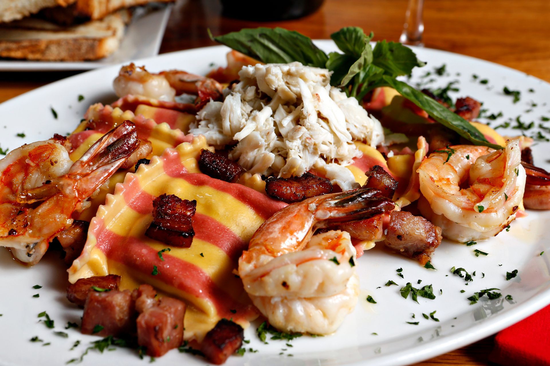 A white platter of striped ravioli topped with crab, shrimp, and bacon pieces, garnished with fresh basil.
