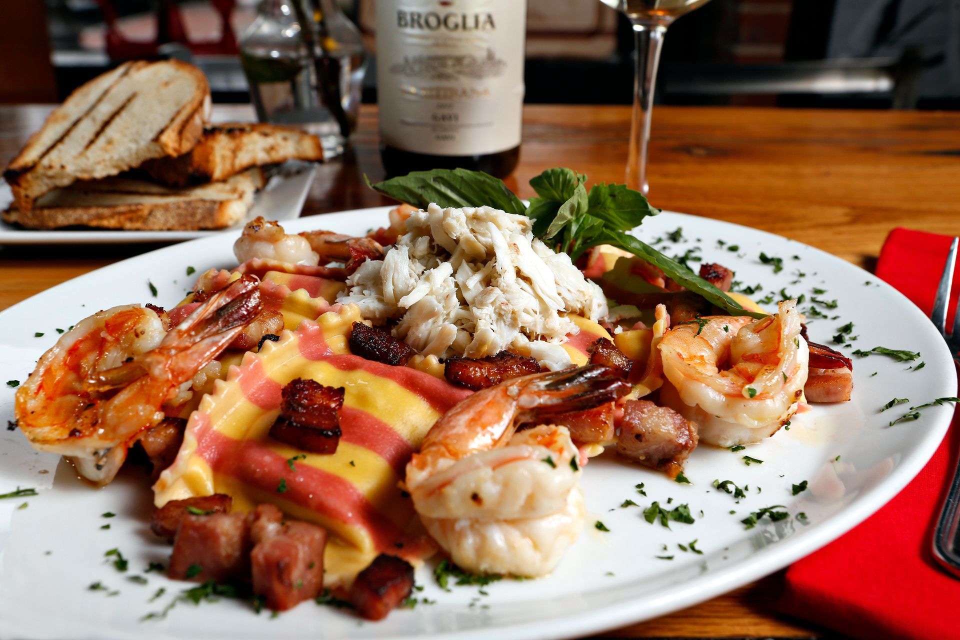A plate of striped ravioli topped with crab meat, shrimp, and bacon pieces, served with toasted bread in a restaurant.