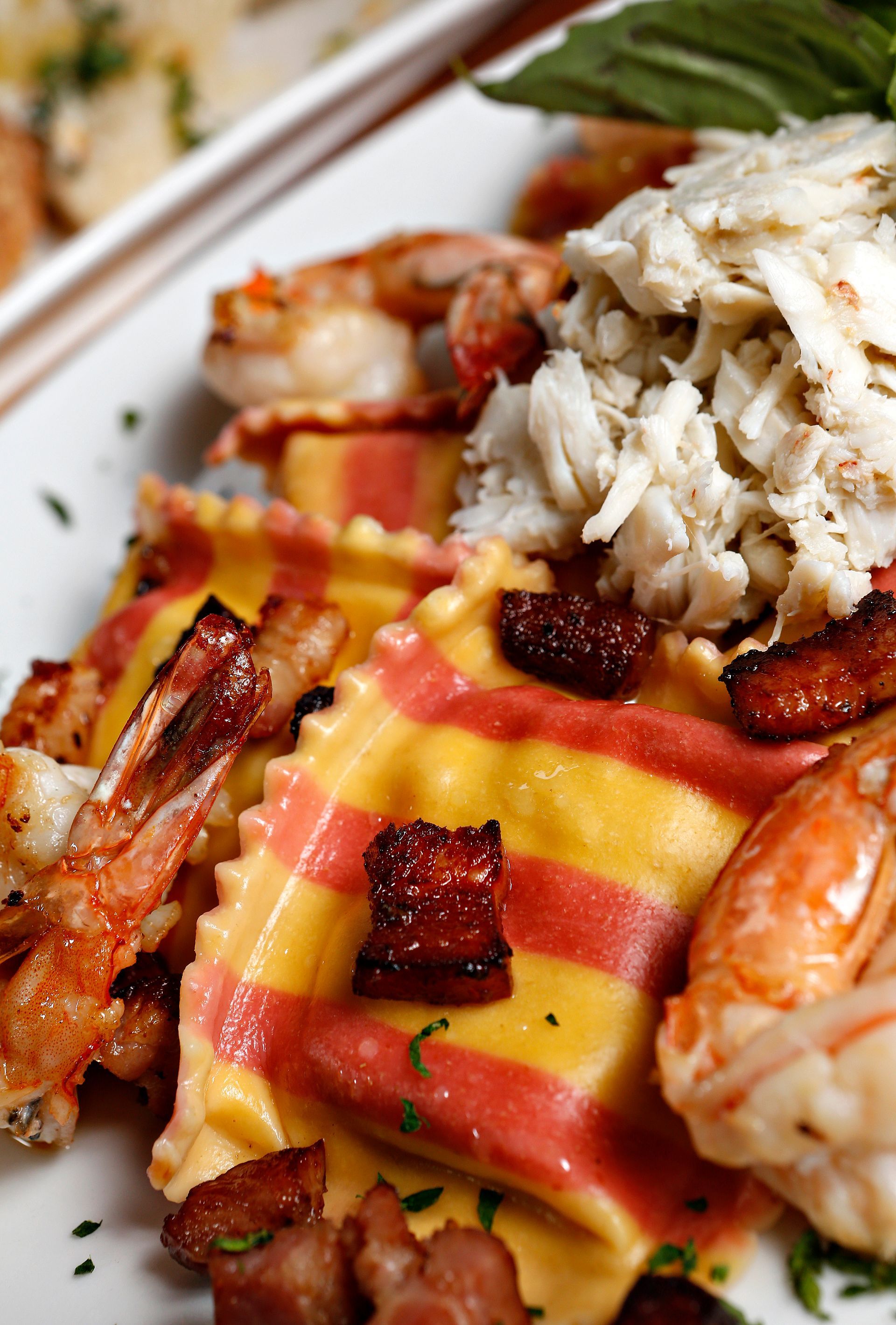 A close-up of striped ravioli served with large shrimp, shredded crab meat, and crispy bacon pieces.