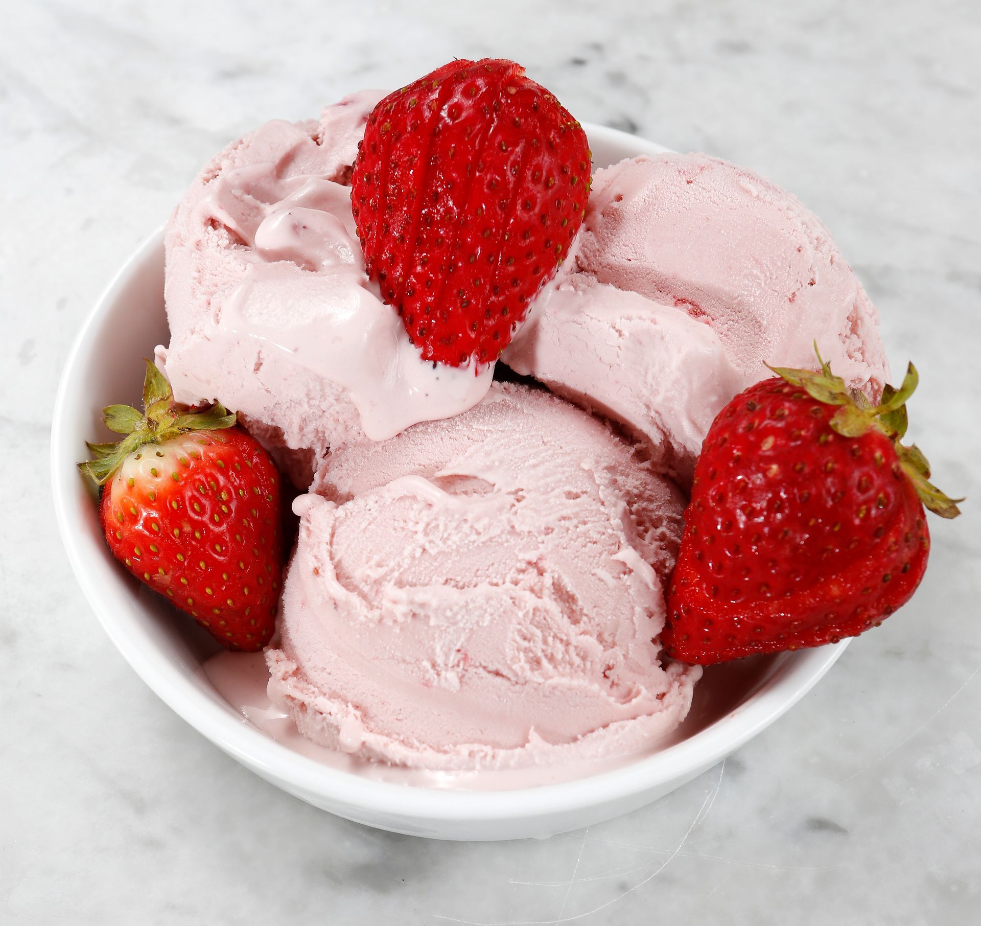 Three scoops of pink strawberry ice cream in a white bowl, garnished with three fresh whole strawberries.