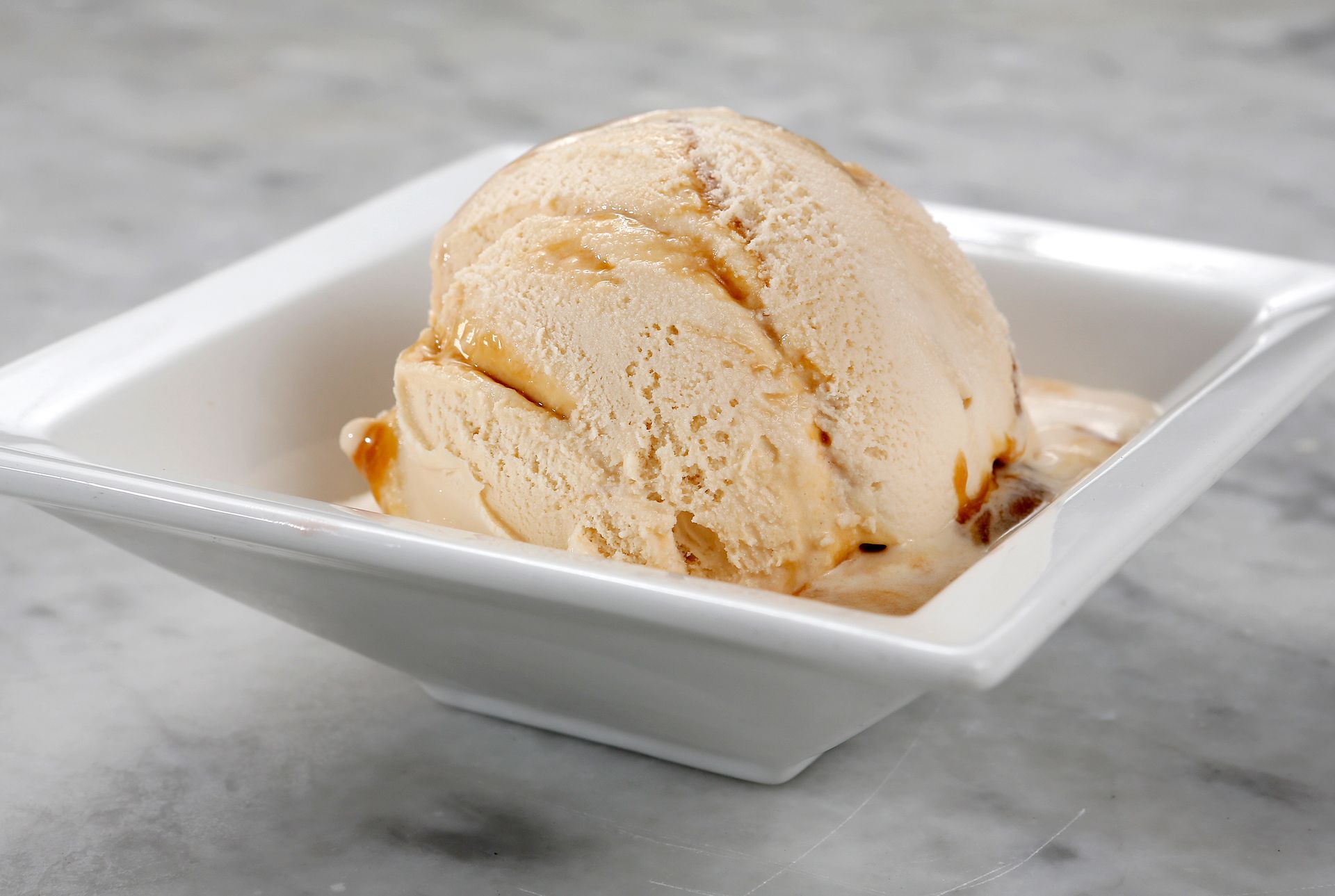 A scoop of caramel swirl ice cream served in a square white bowl on a marble surface.