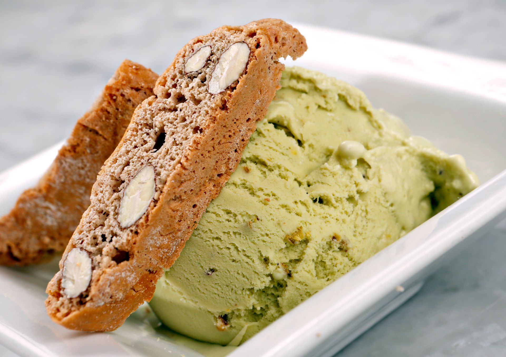 A scoop of light green pistachio ice cream served in a white bowl with two almond biscotti cookies.