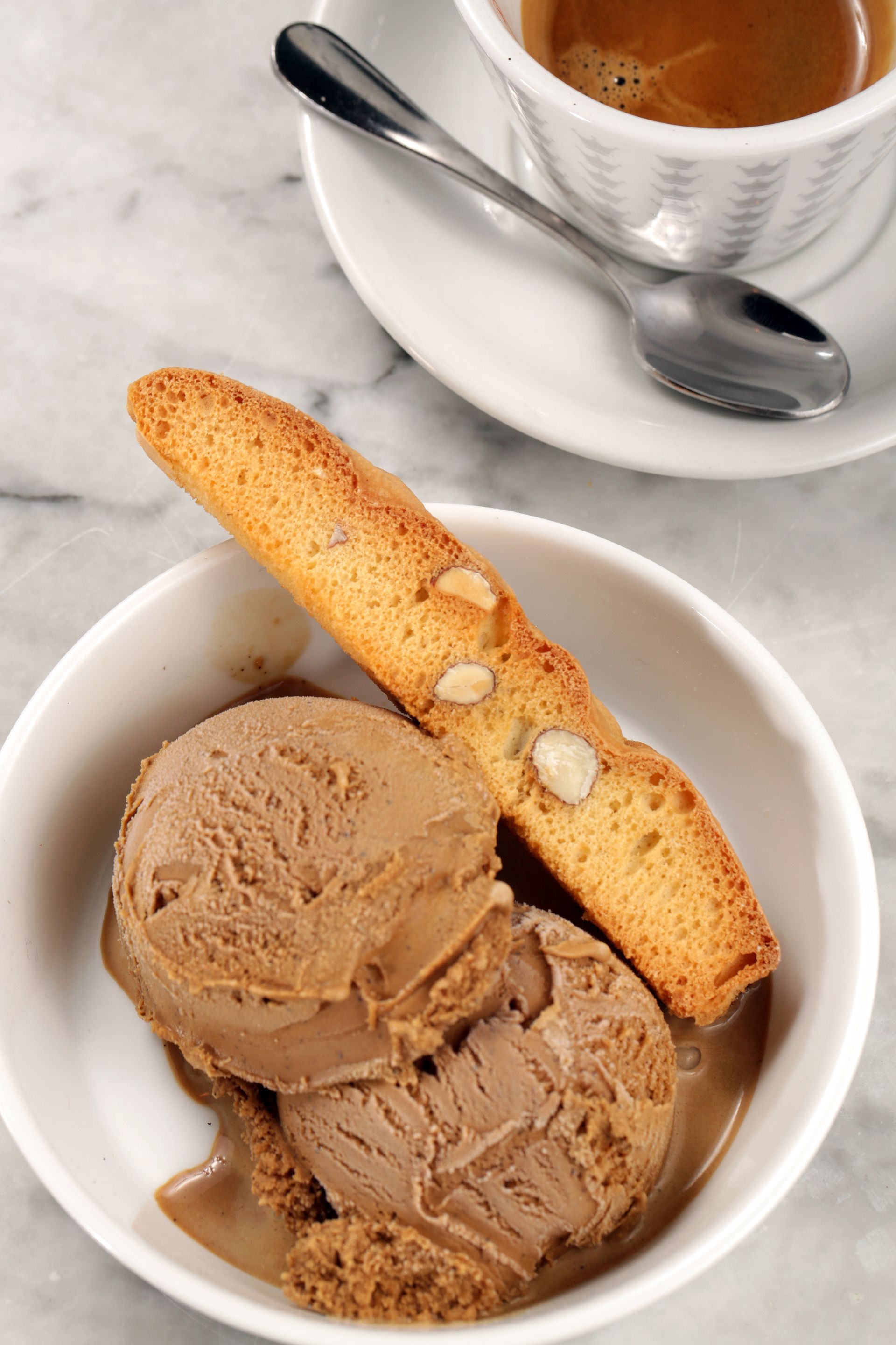 A bowl of two scoops of chocolate gelato with a biscotti, next to a small cup of espresso on a marble surface.