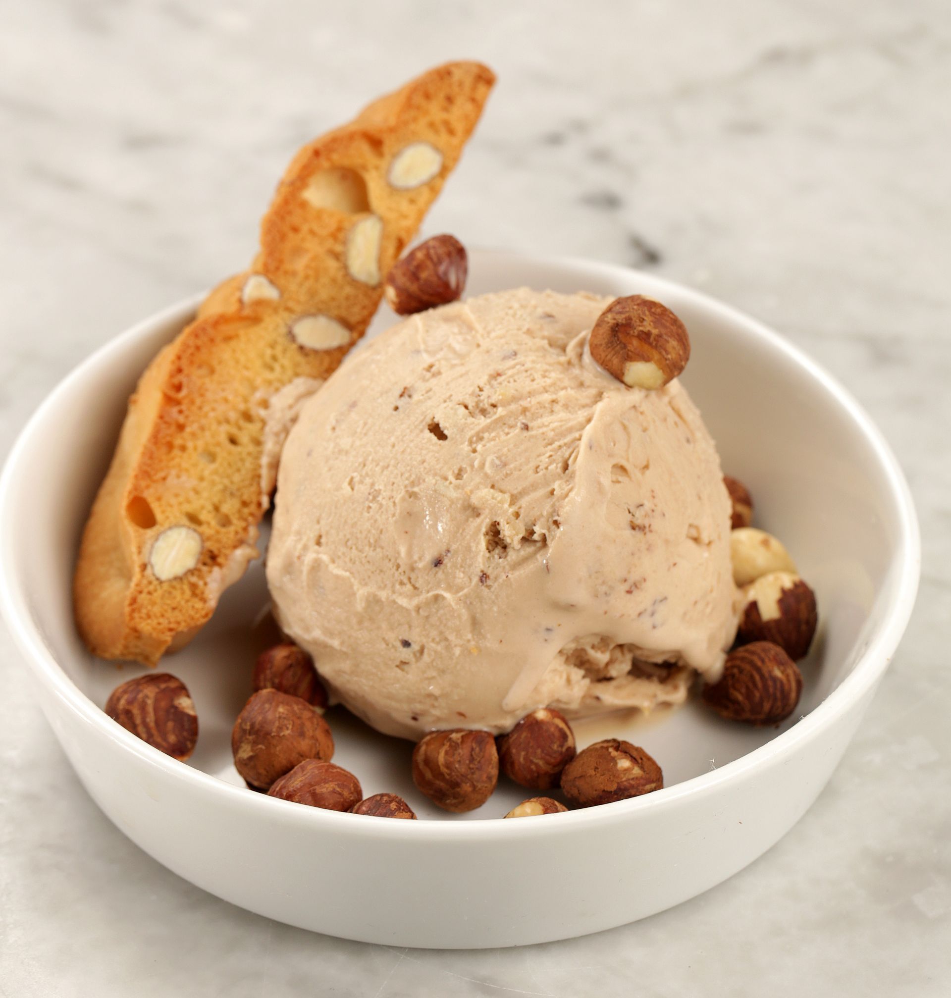 A scoop of light brown hazelnut ice cream in a white bowl, garnished with whole hazelnuts and a piece of biscotti.