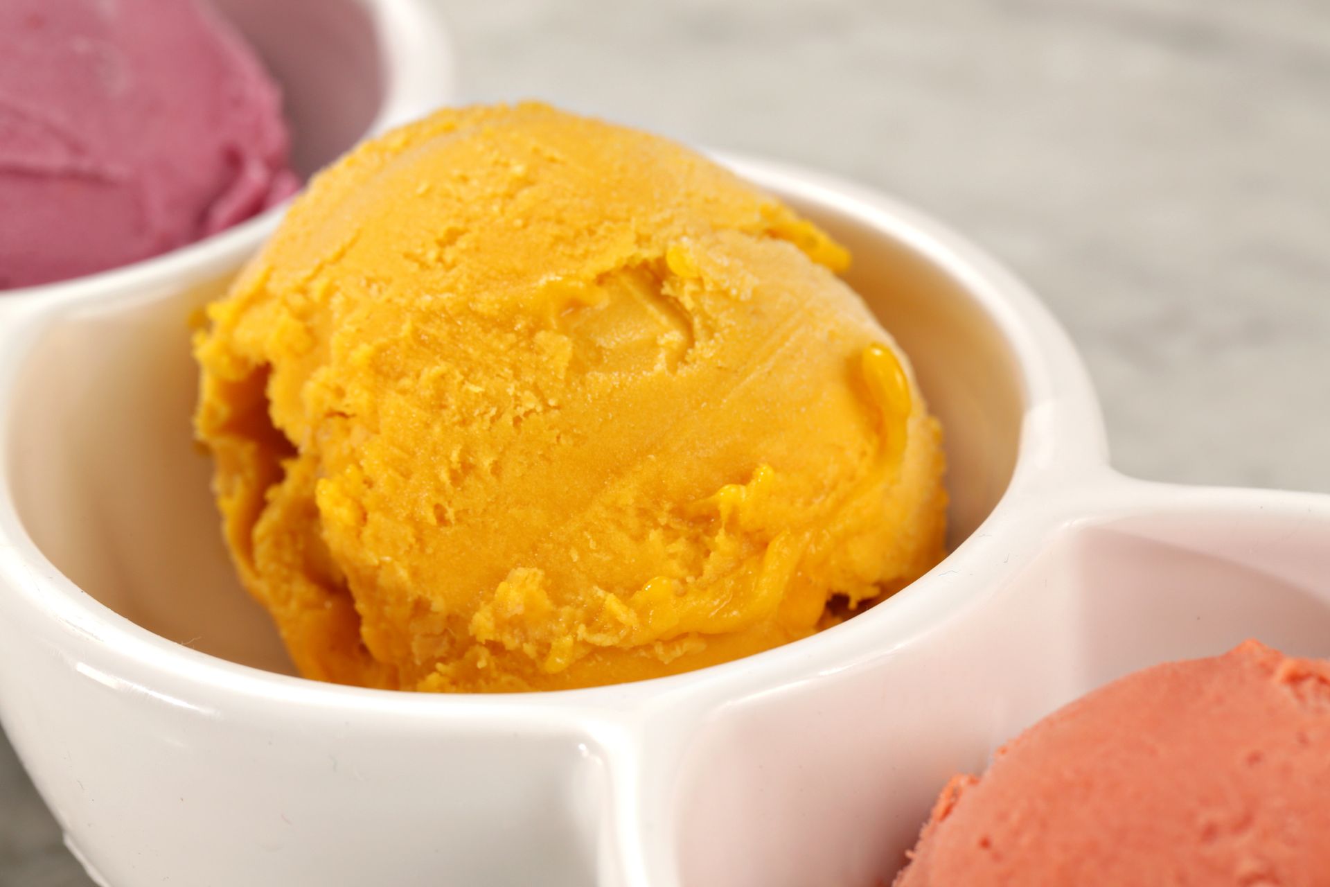 A close-up of a bright orange scoop of ice cream centered in a white, partitioned serving dish with other flavors.