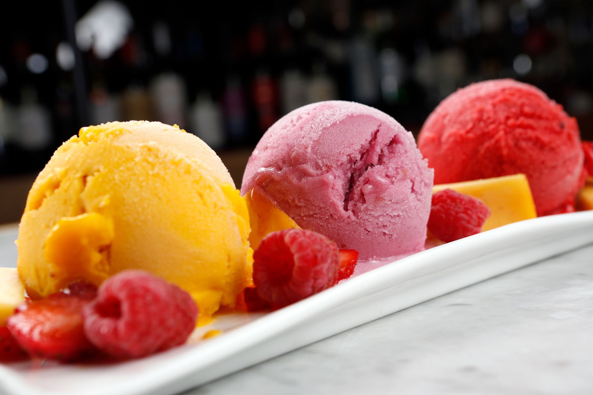 Three scoops of sorbet—yellow, pink, and red—on a rectangular white plate, garnished with fresh raspberries and strawberries.
