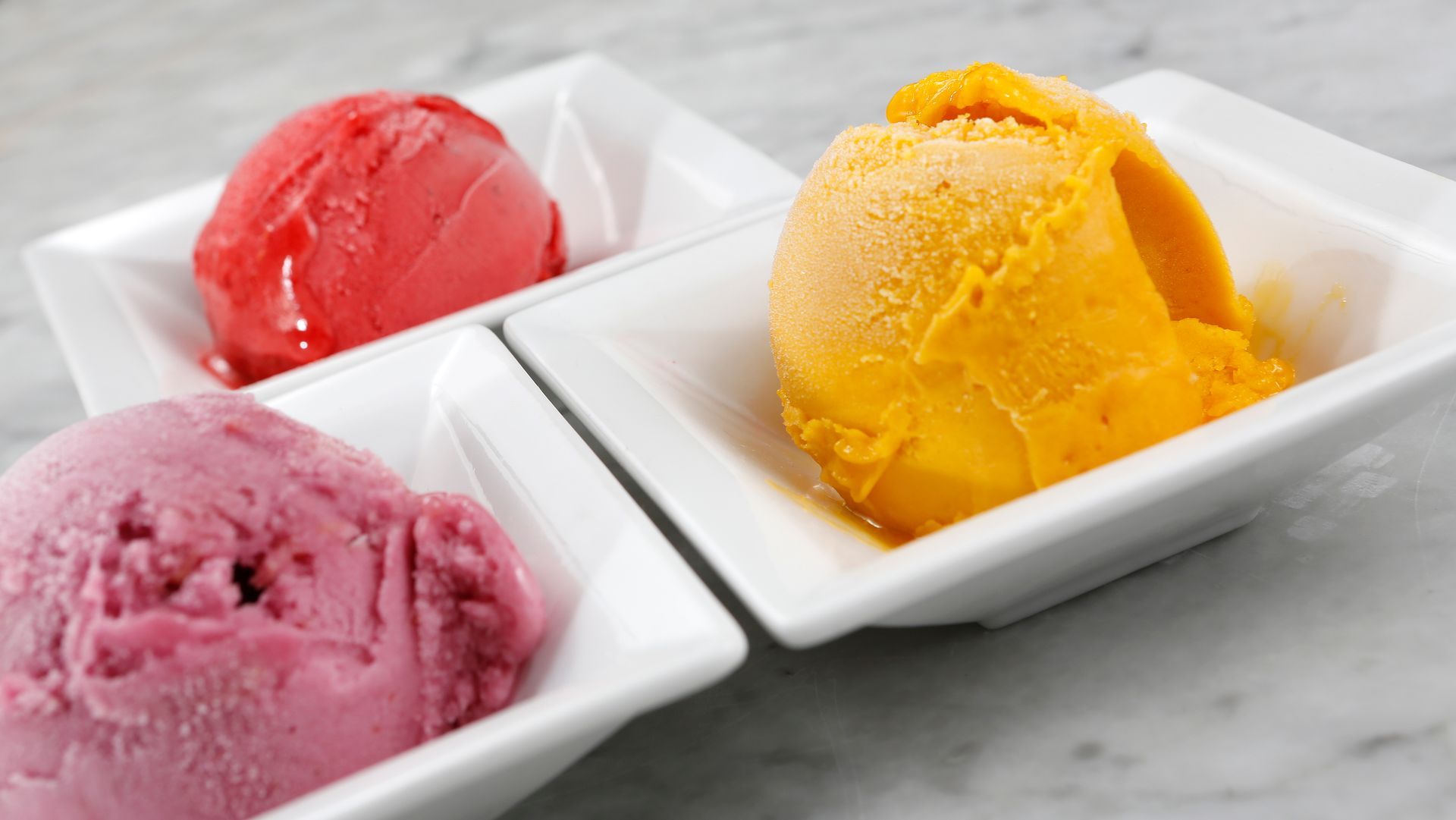 Three scoops of fruit sorbet in individual white square bowls on a marble surface, colored red, purple, and orange.