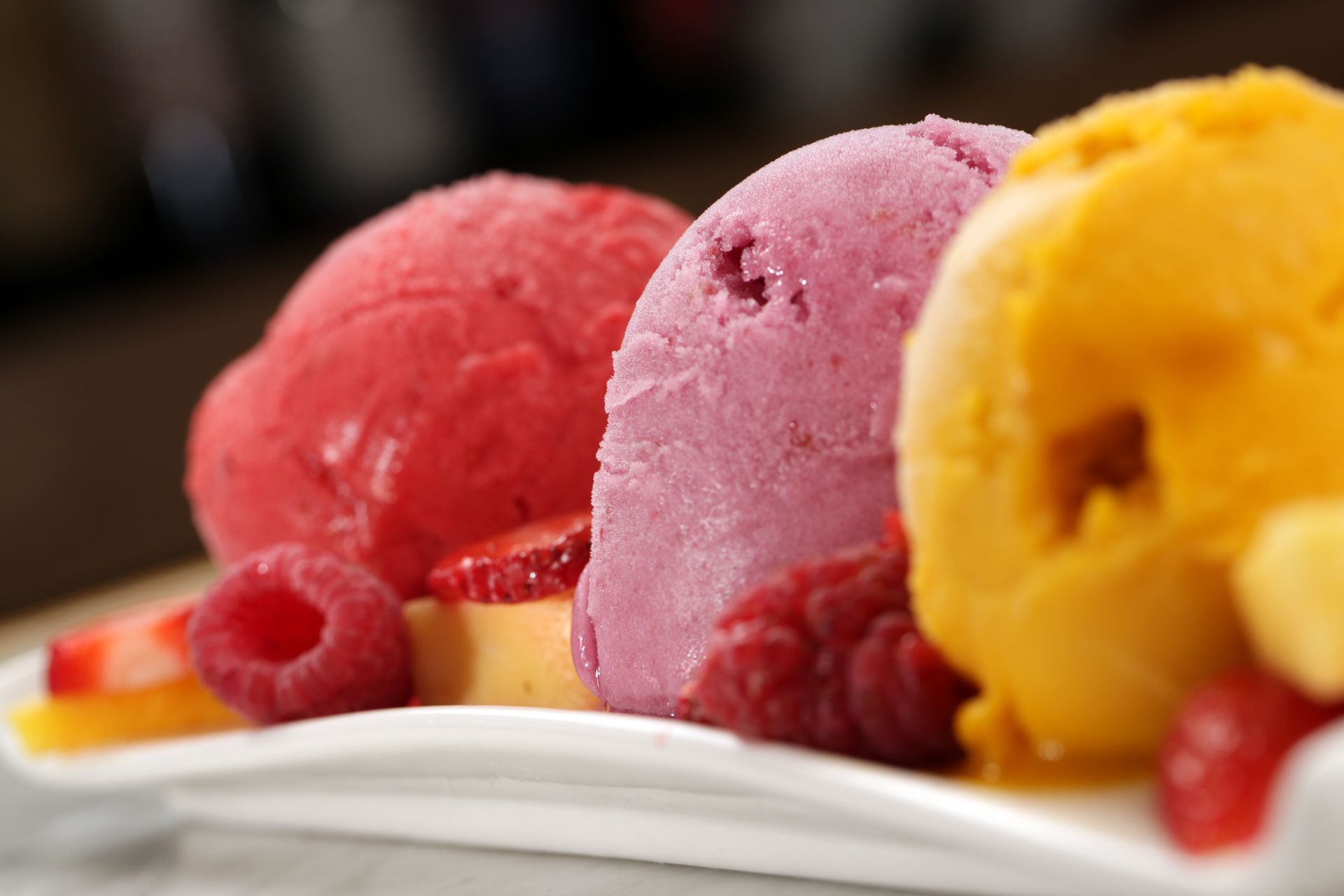 Three scoops of red, purple, and yellow sorbet garnished with fresh raspberries and sliced strawberries on a white plate.