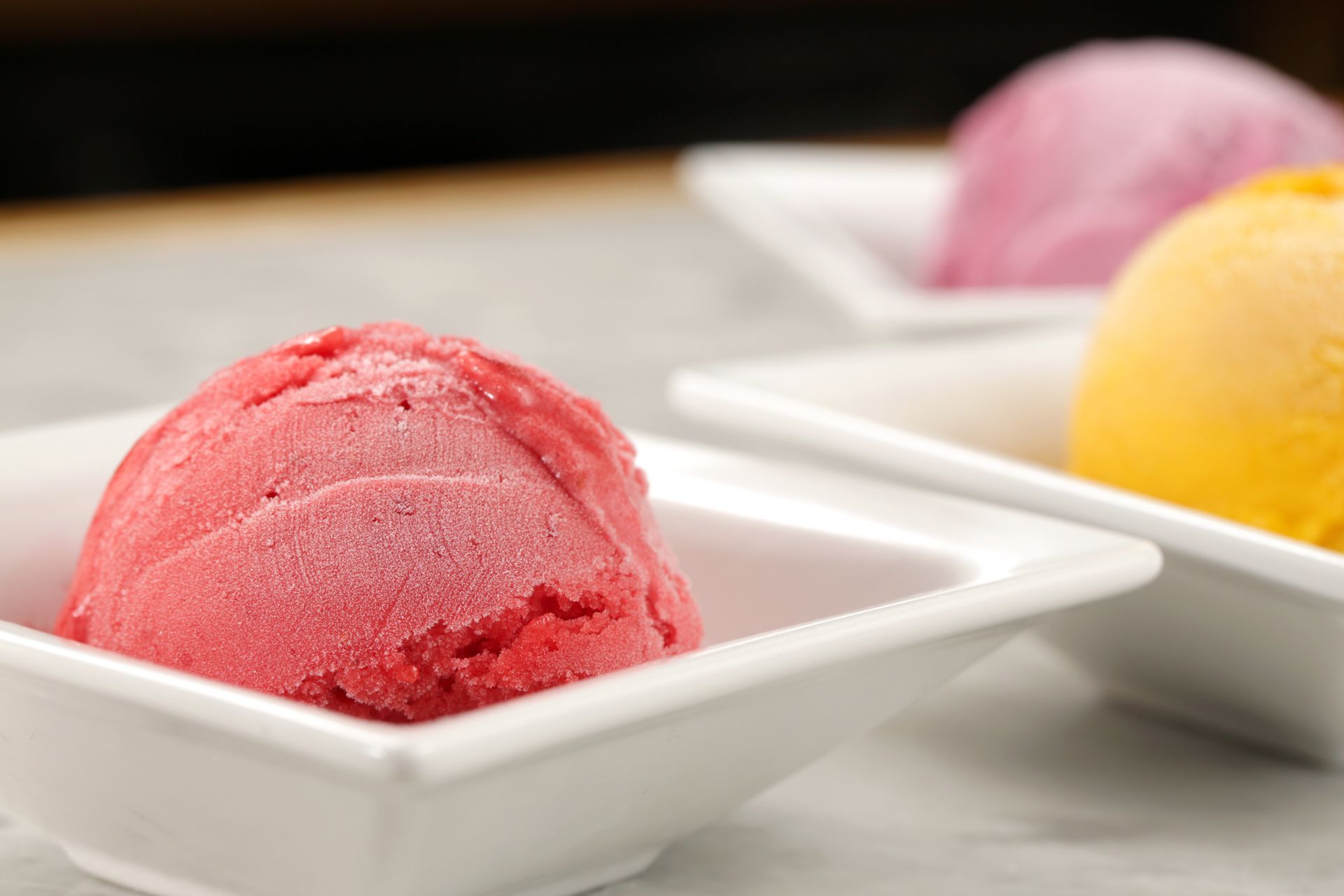 Three scoops of pink, yellow, and red sorbet or ice cream served in individual white bowls on a light surface.