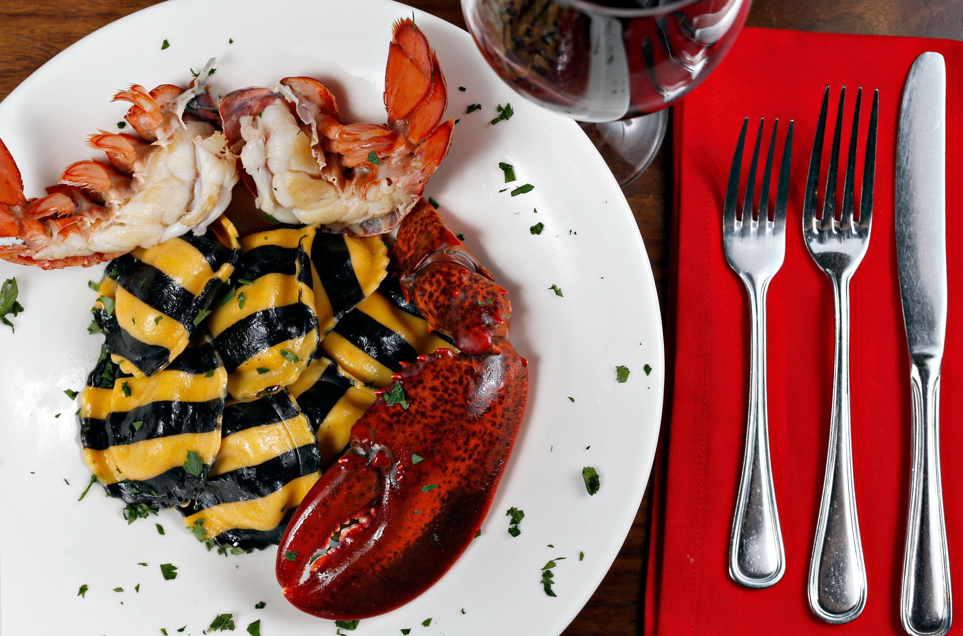 A plate of striped black and yellow ravioli with two lobster tails, next to a glass of wine and silverware on a red napkin.