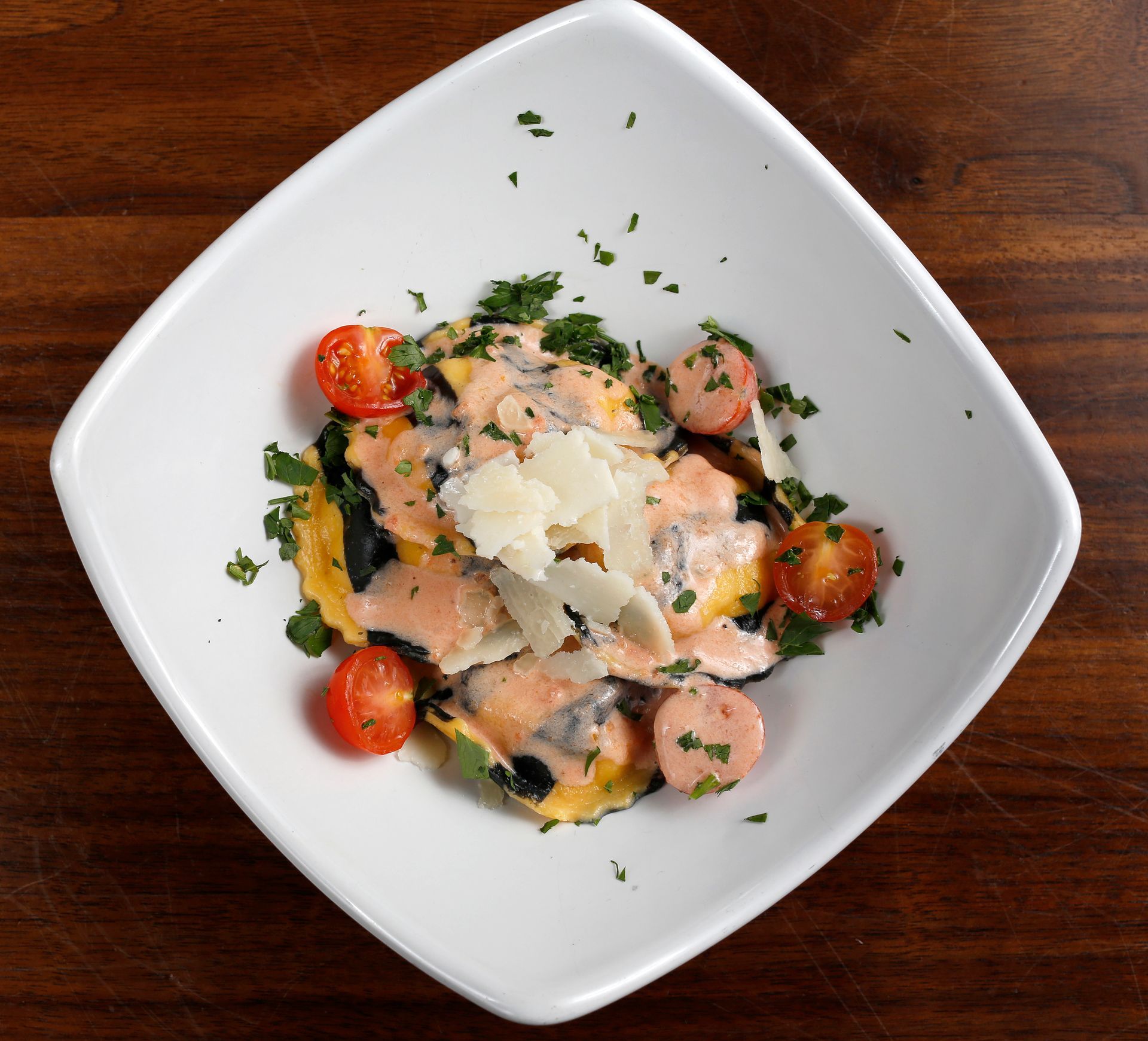 A white bowl containing ravioli in a creamy pink sauce, topped with shaved cheese, cherry tomatoes, and fresh herbs.