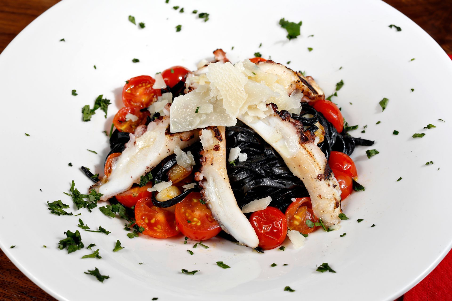 A serving of black squid ink pasta topped with grilled octopus, cherry tomatoes, and shaved parmesan on a white plate.