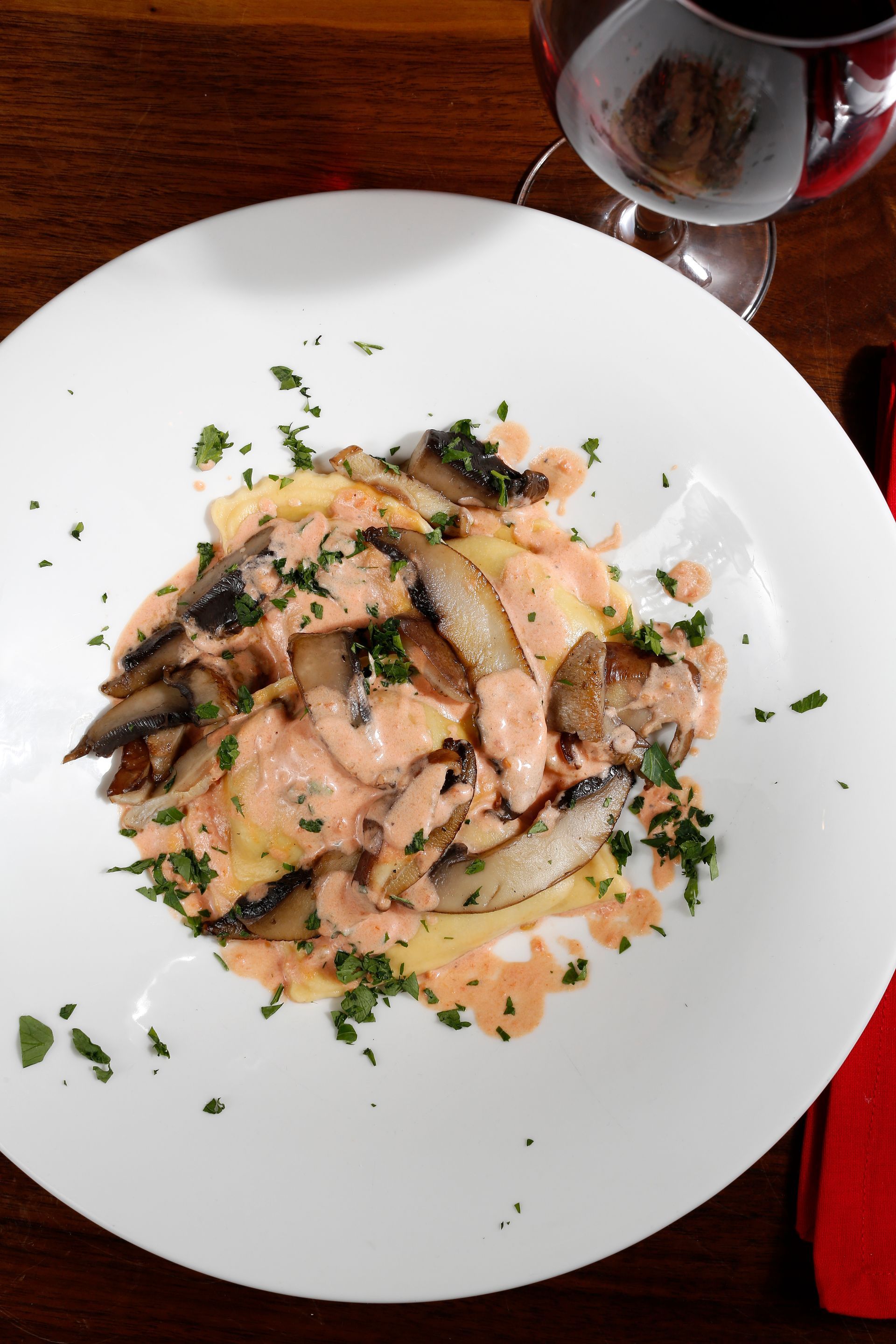 A plate of mushroom ravioli topped with a creamy pink sauce and fresh herbs, next to a glass of red wine.