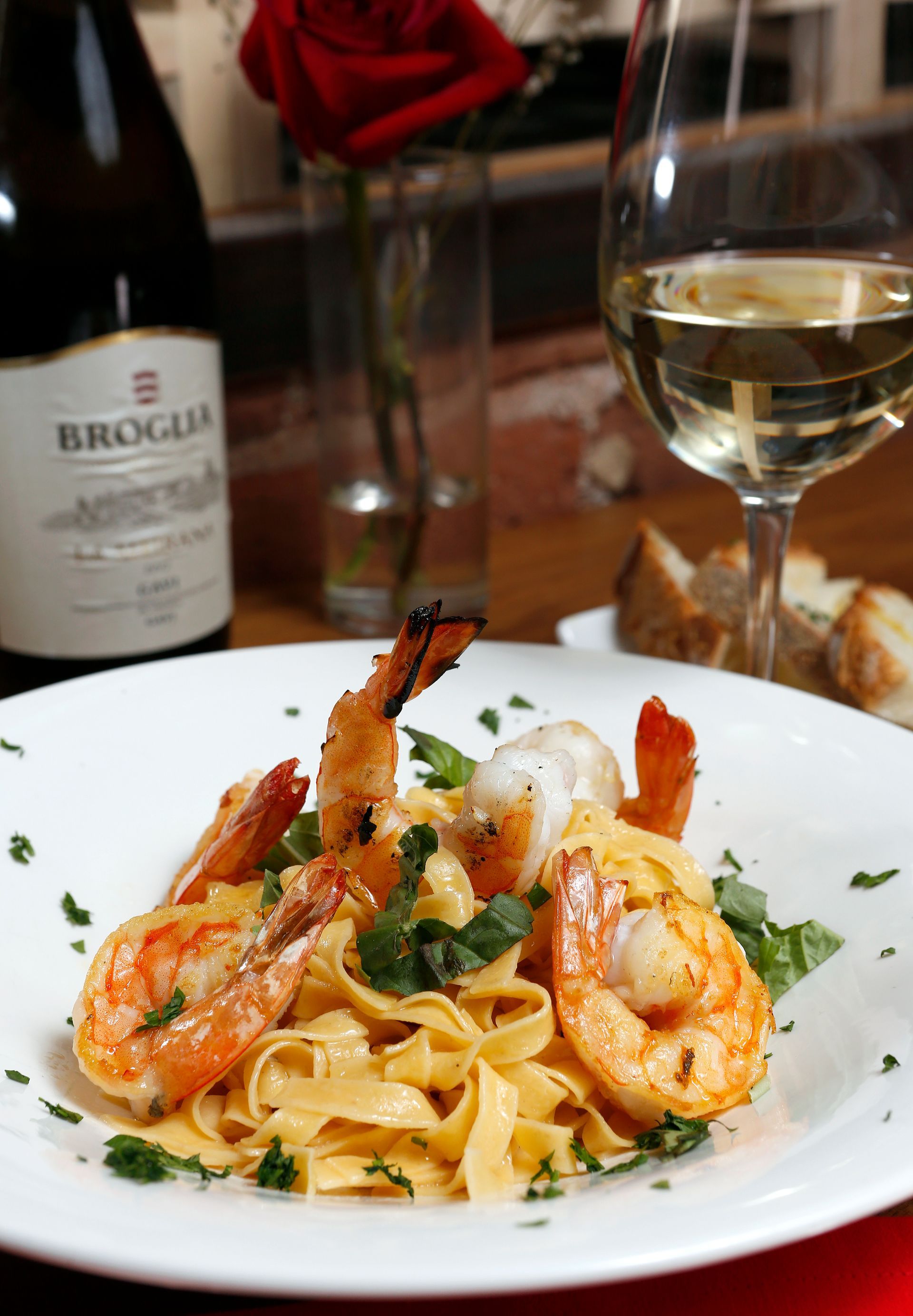 A white bowl of shrimp fettuccine garnished with herbs, with a glass of white wine and a wine bottle in the background.