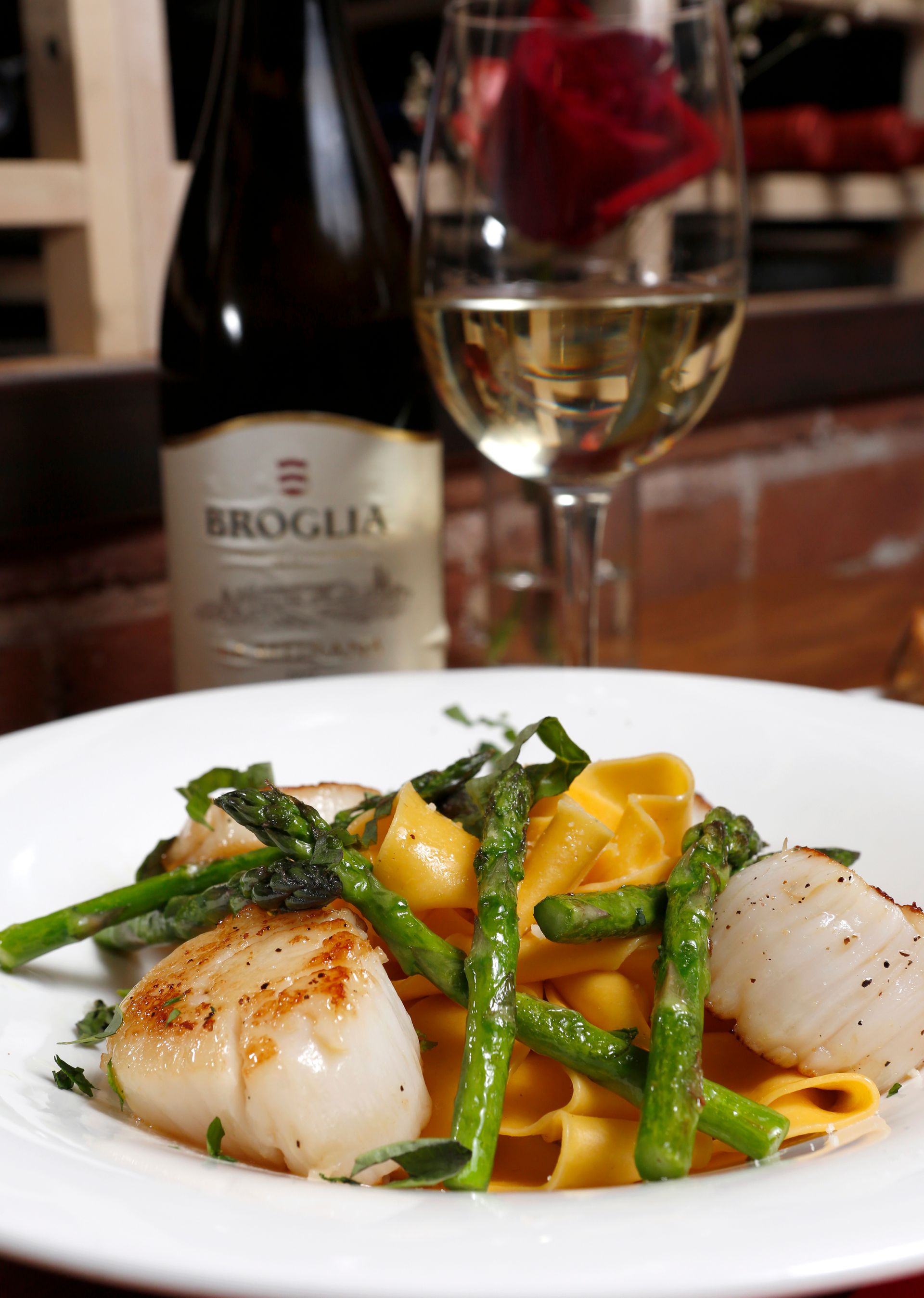 A white plate of seared scallops and asparagus over golden pasta, with a glass of white wine and a Broglia bottle behind.
