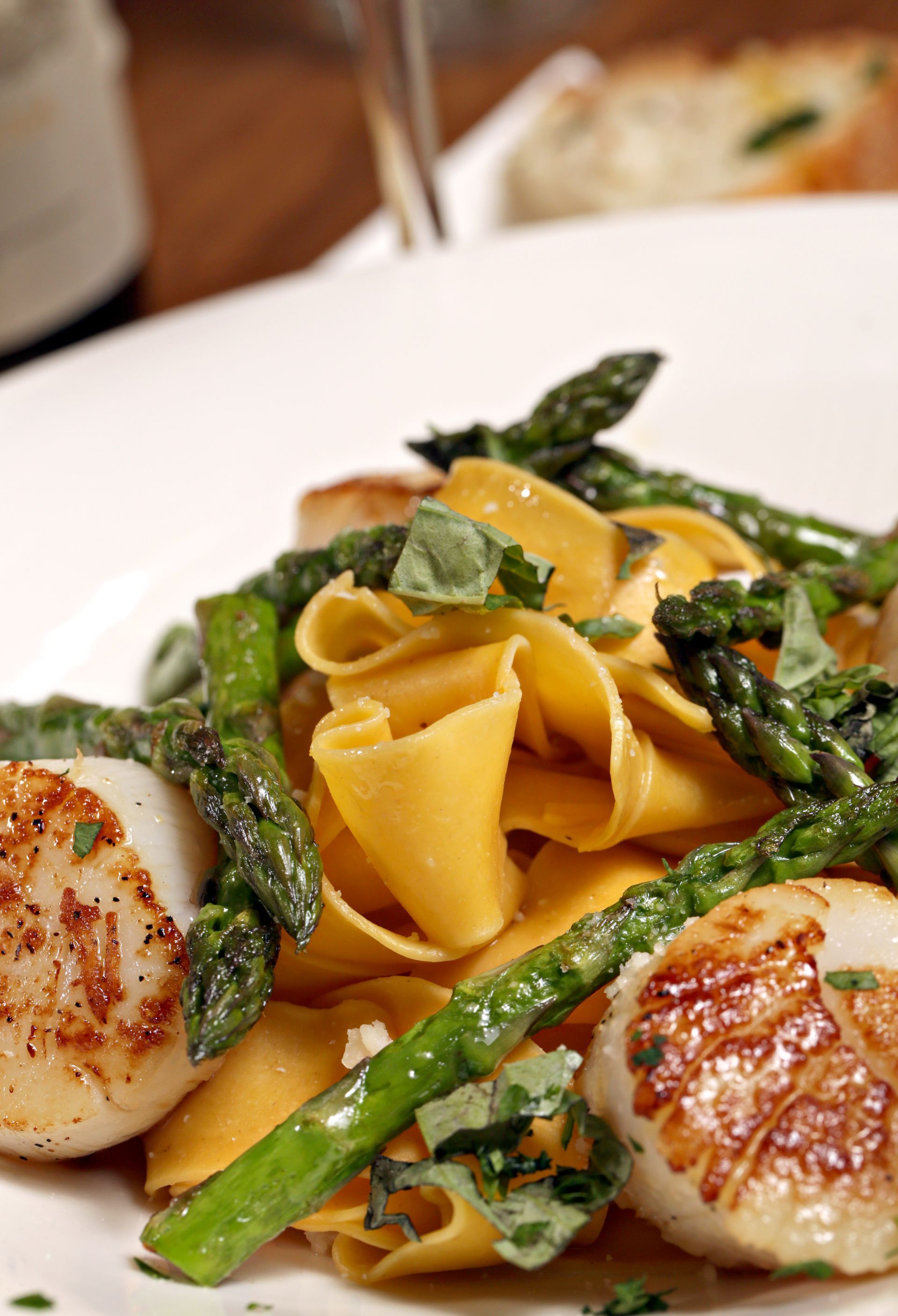 A white plate holds seared scallops served over wide pasta ribbons with asparagus spears and fresh green herbs.