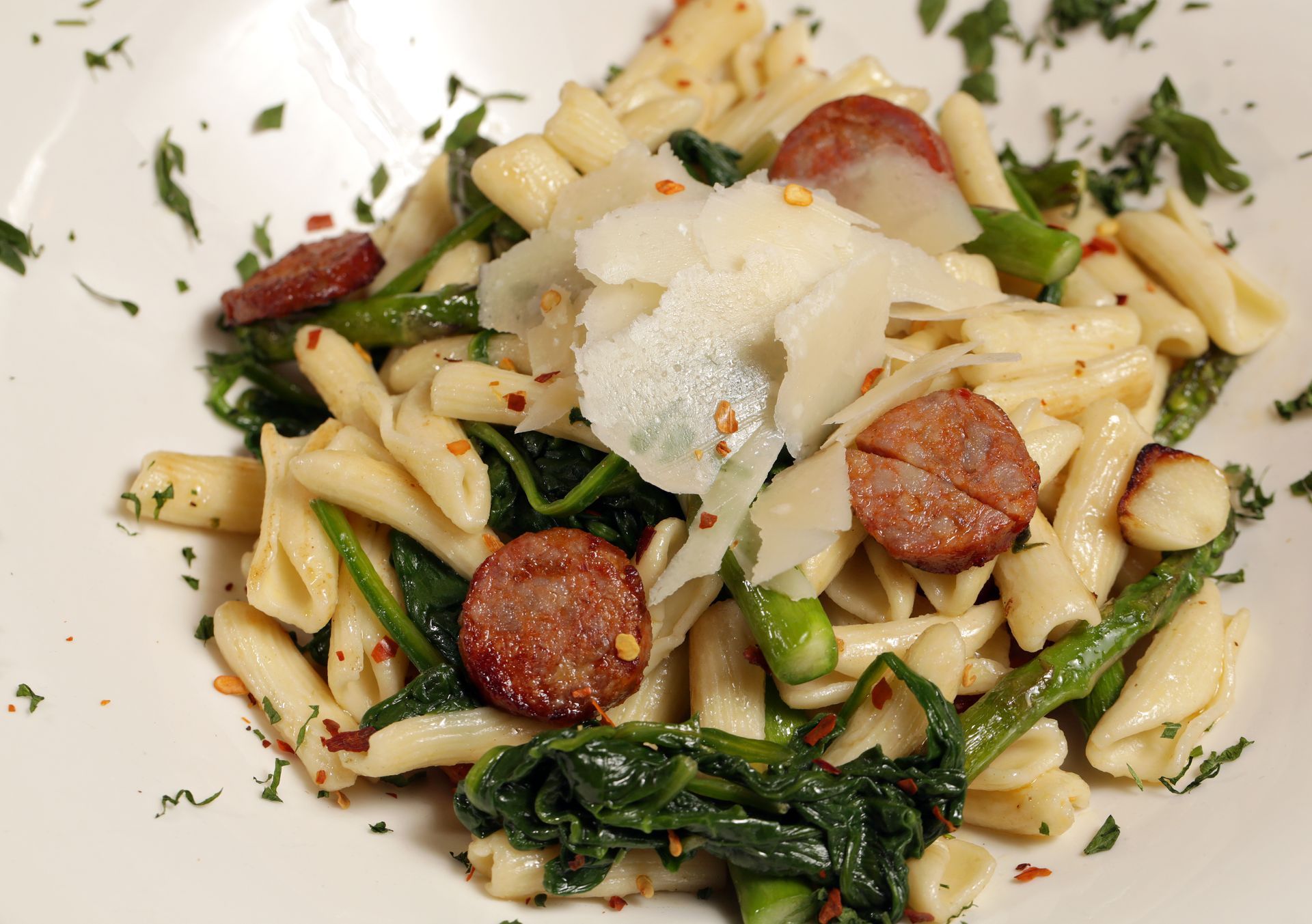 A bowl of pasta with sliced sausage, sautéed greens, asparagus, and shaved cheese topped with red pepper flakes.