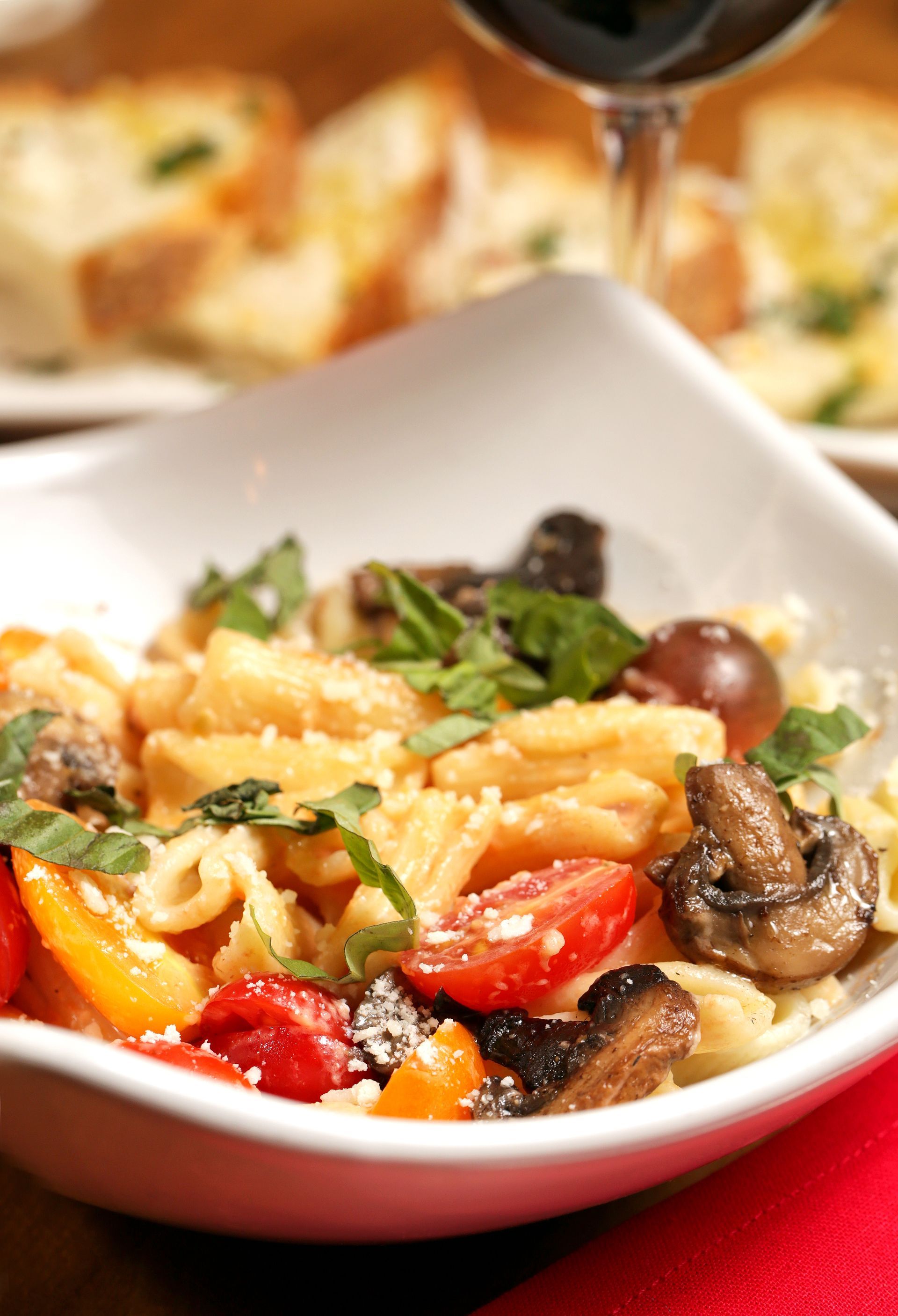 A white bowl of pasta with mushrooms, cherry tomatoes, and fresh basil, served with bread and red wine in the background.