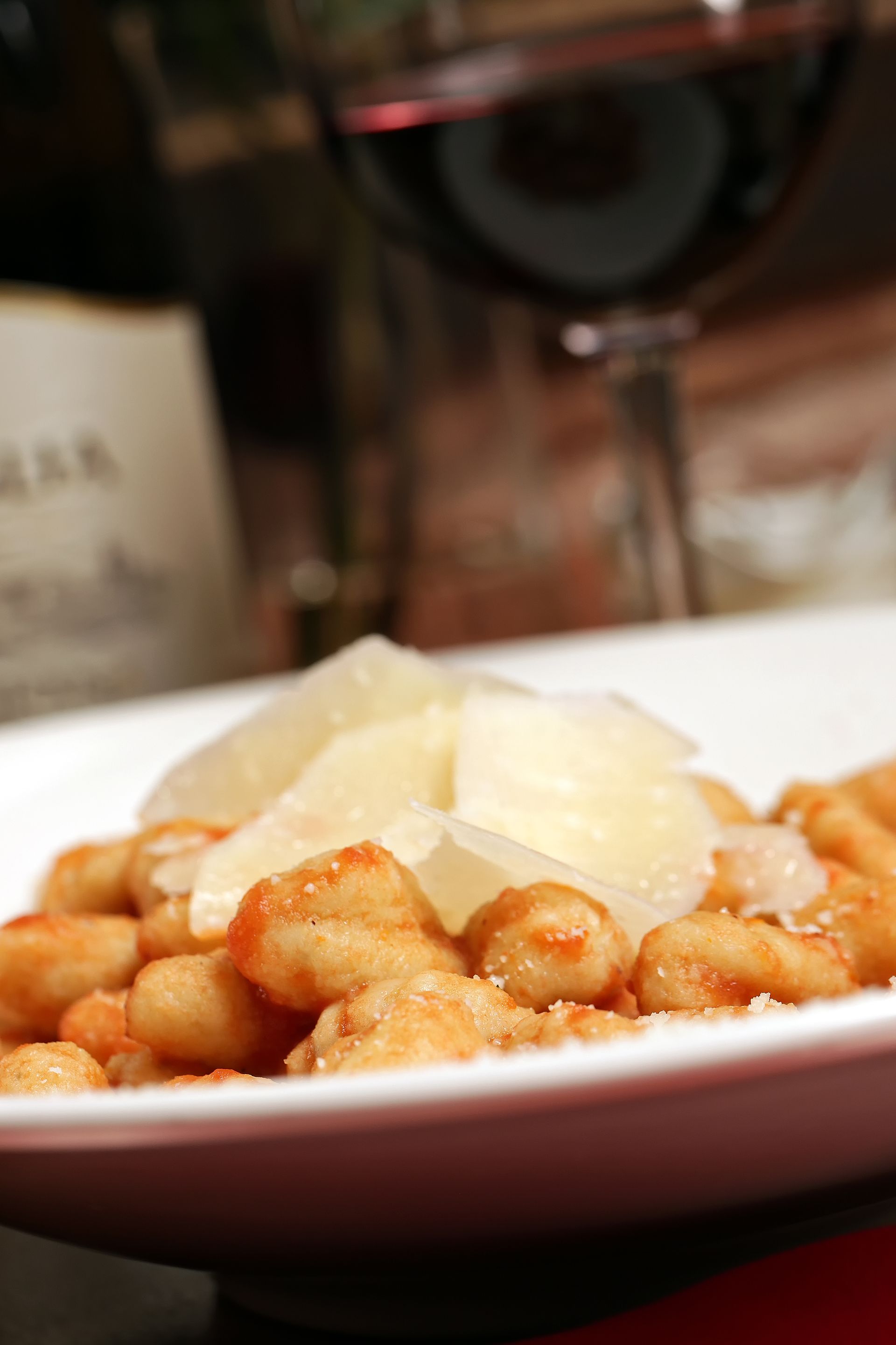 A plate of gnocchi topped with shaved parmesan cheese, with a blurred wine bottle and glass in the background.