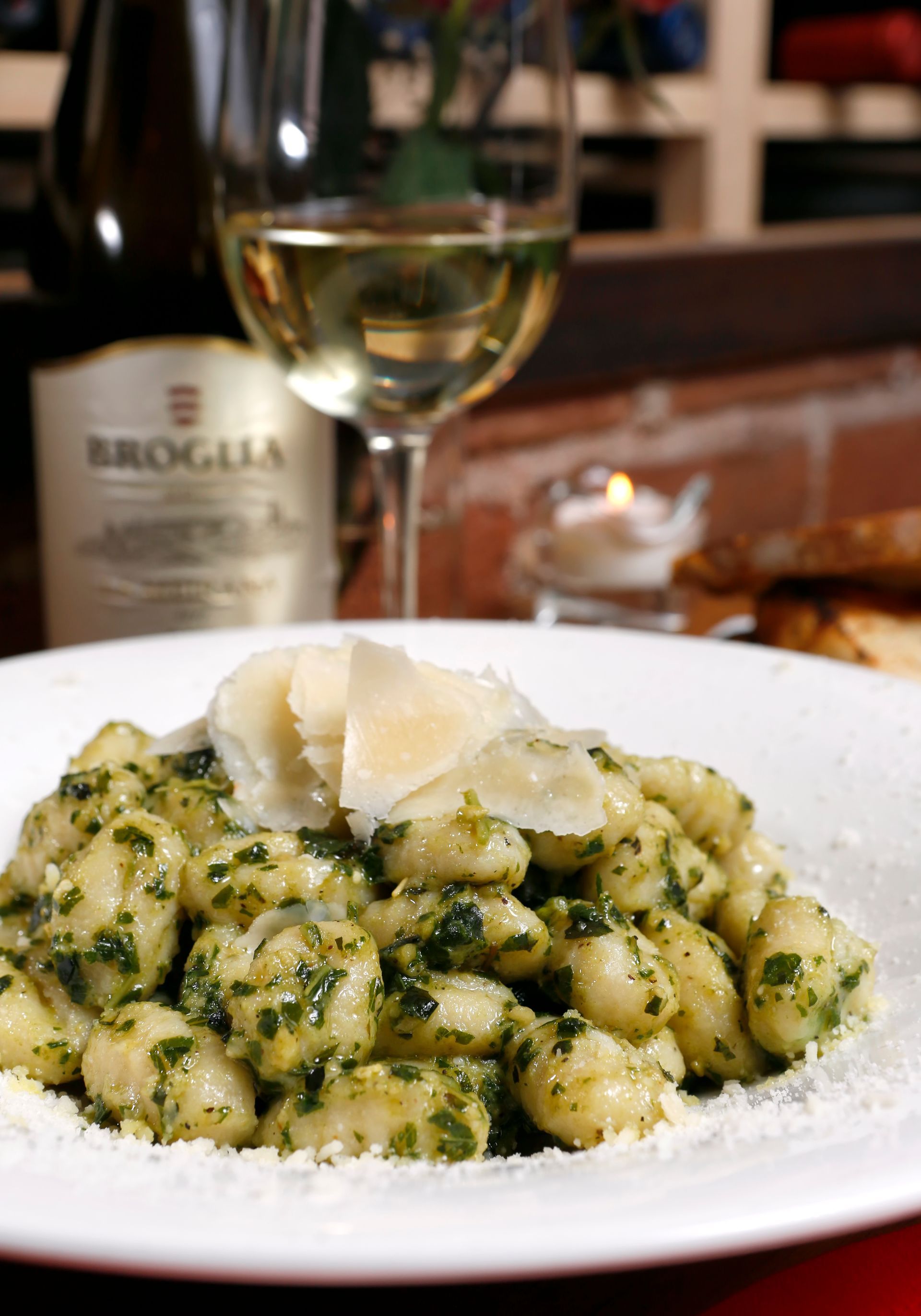 A plate of pesto gnocchi topped with parmesan shavings, with a glass of white wine and a bottle of Broglia in the back.