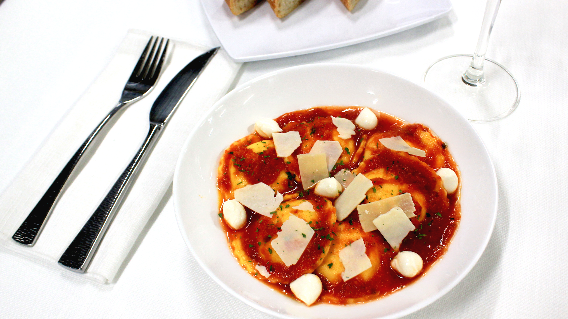 A white bowl of ravioli in red sauce with mozzarella balls and parmesan shavings, next to cutlery and bread on a table.