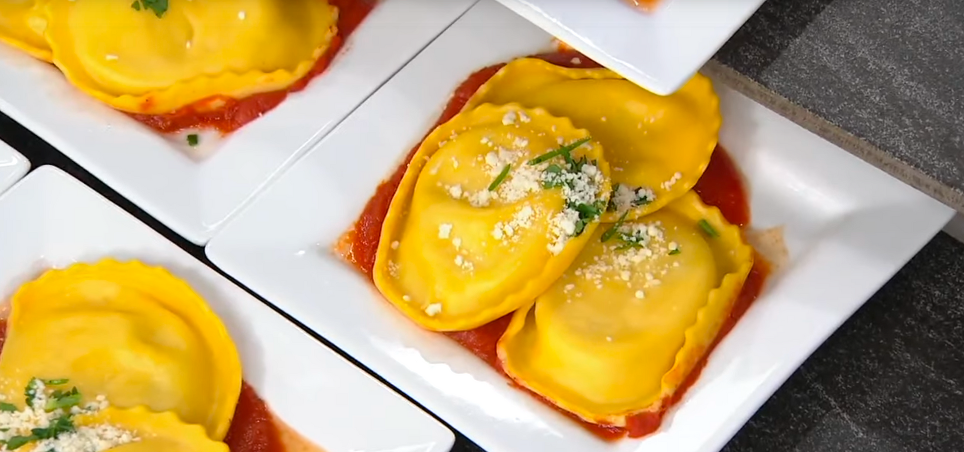 Three square white plates holding bright yellow ravioli in red tomato sauce, topped with grated cheese and herbs.