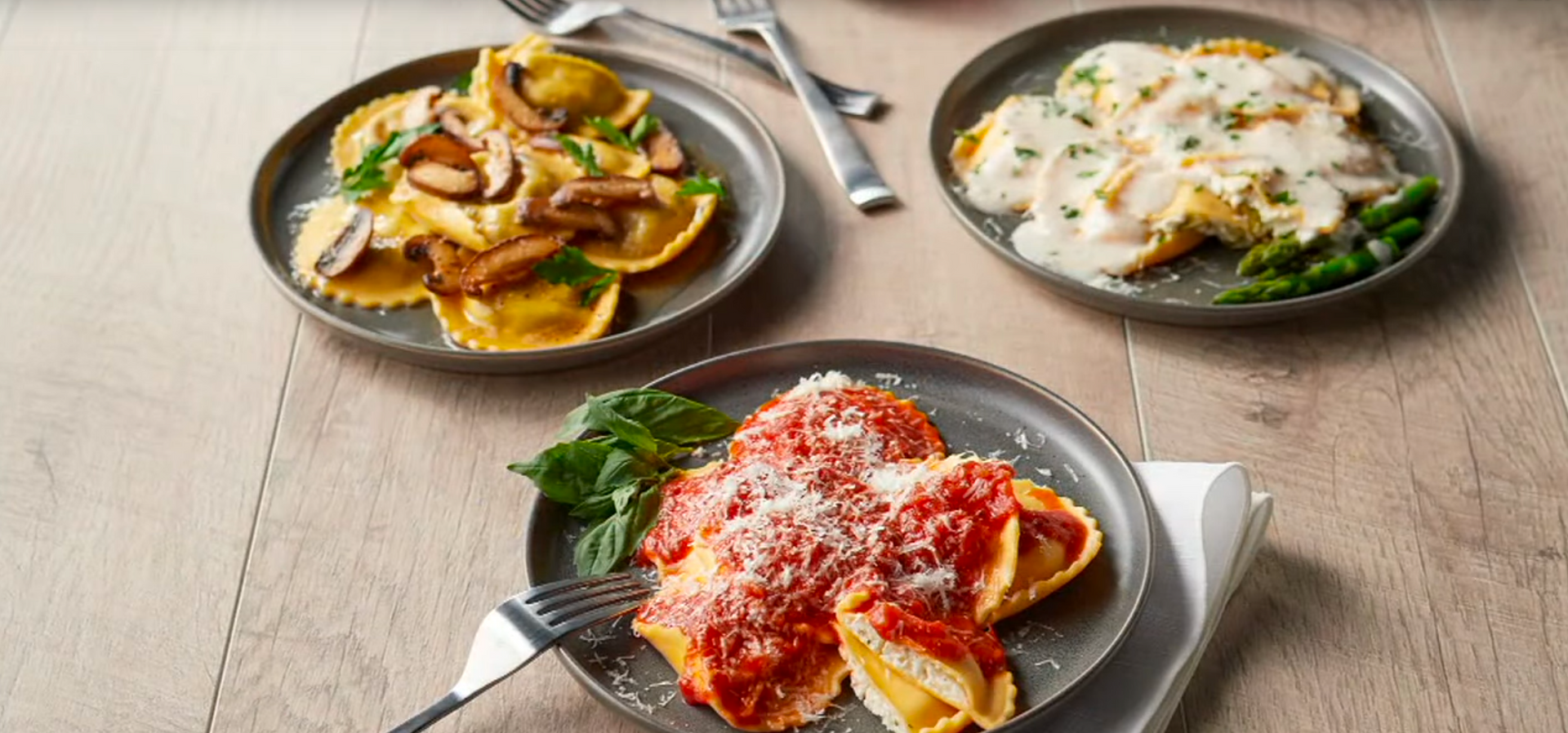 Three plates of ravioli on a wooden table, served with mushroom, cream, and tomato sauce, with forks nearby.