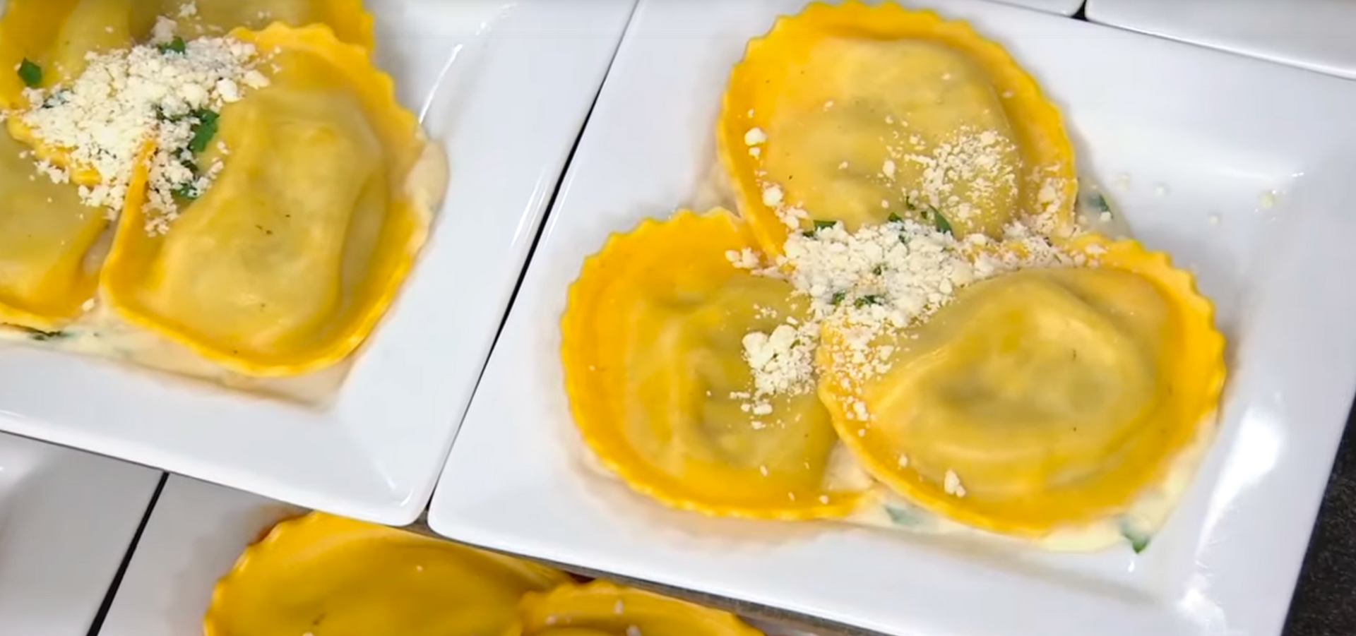 Two white plates hold golden, circular ravioli topped with white cheese and green herbs, served in a creamy sauce.