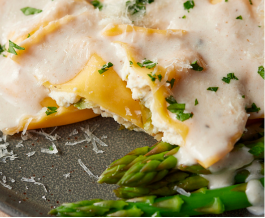 Ravioli filled with white cheese, topped with a creamy white sauce and parsley, served alongside fresh green asparagus.