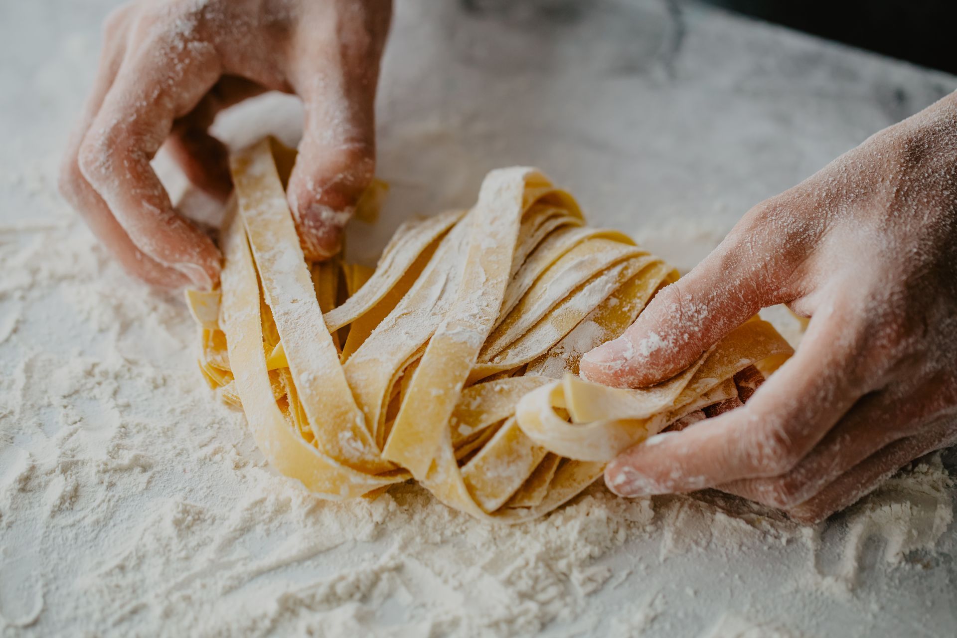 Hands dusted in flour gently shape fresh, golden tagliatelle pasta on a floured surface.