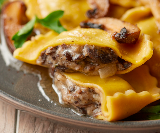 A close-up of mushroom-stuffed ravioli garnished with sautéed mushroom slices and fresh herbs on a dark plate.