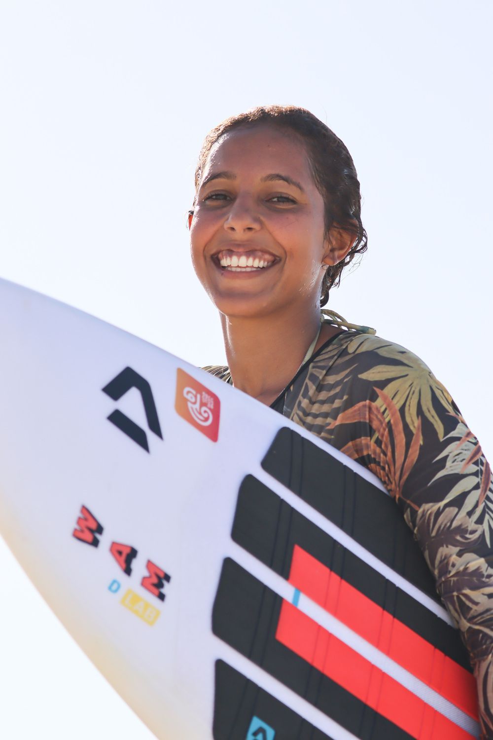 Uma mulher sorri enquanto segura uma prancha de surfe com a inscrição b4z