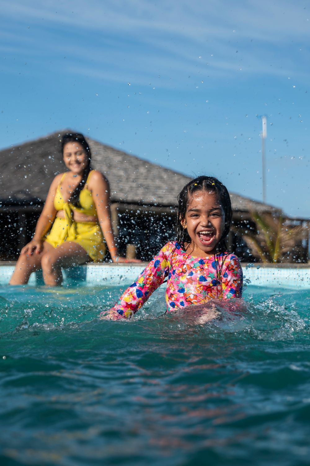 Uma mulher está sentada na beira de uma piscina enquanto uma menina brinca na água.