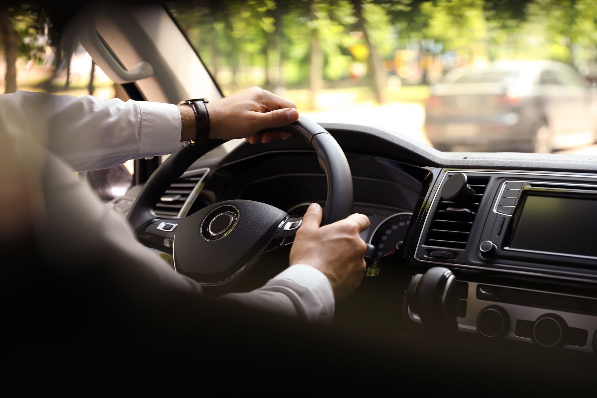 Driver’s hands on a car steering wheel, driving through a sunlit street.