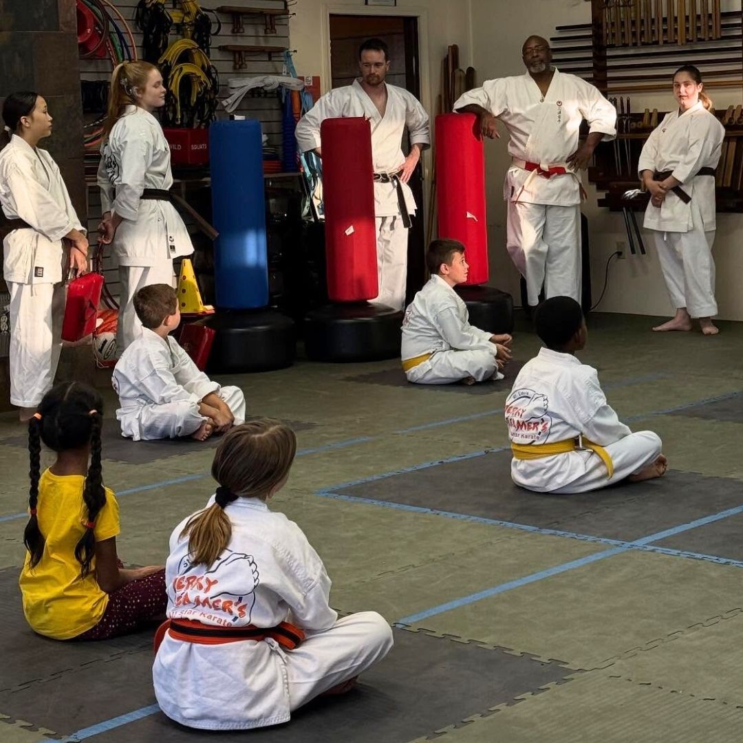 Two young children in blue and black martial arts uniforms grappling on a gym floor, one smiling.