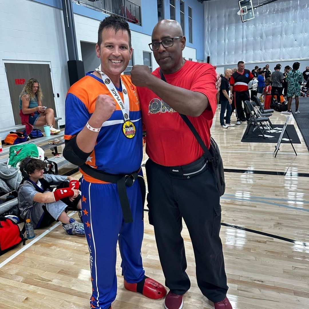 Two men in grappling position on a mat. One embraces the other from behind, both smiling. Gym setting.
