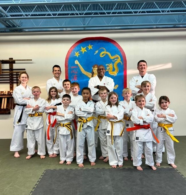 Family in Jiu-Jitsu gis poses. Indoors, blue mats, a smiling woman in the middle, around a seated man with a beard.