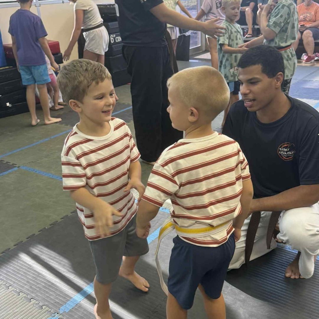 Children in colorful gis play a game on a mat. Two instructors pull a student in blue.