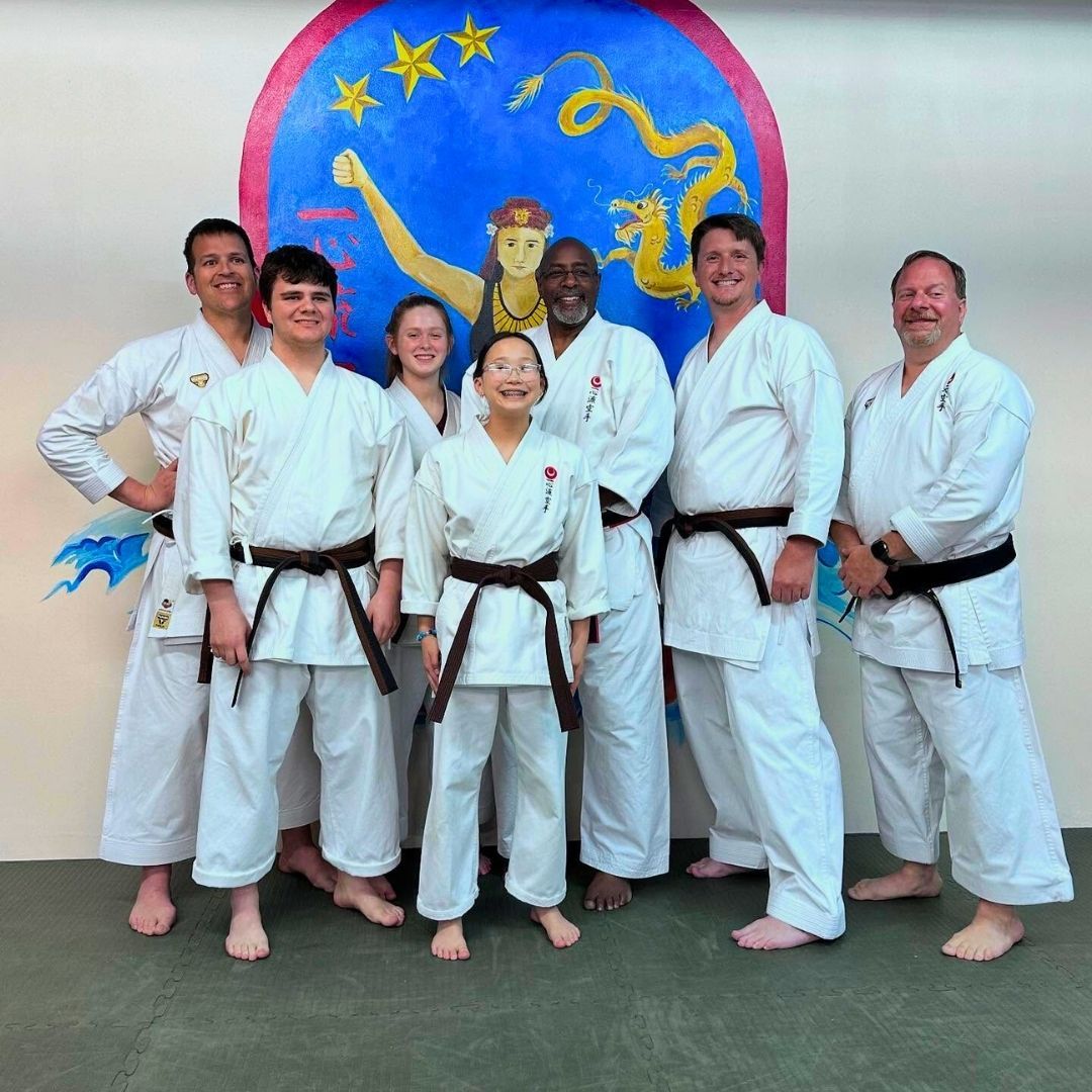 Group of children in martial arts attire posing, some with arms around each other, in a gym.