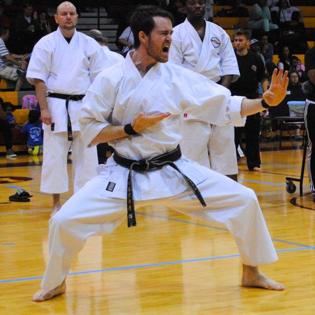 Five people in jiu-jitsu gear pose together in a gym. Smiling, some have arms around each other. Machado logo visible.