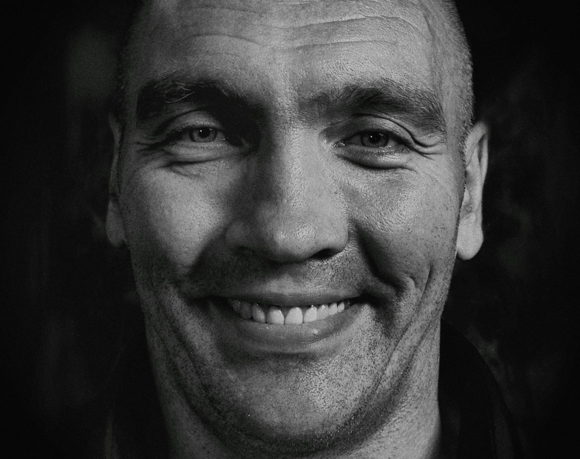 Close-up black and white portrait of a man smiling, with a shaved head and visible teeth.