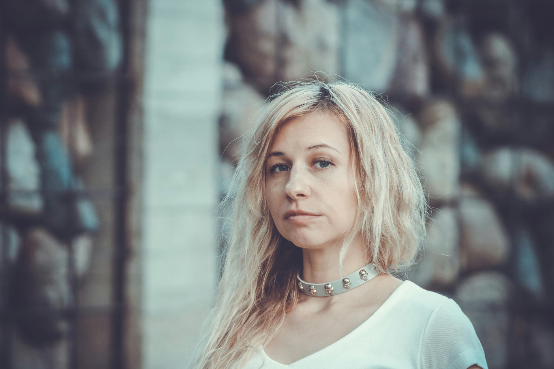 Blond woman wearing a studded choker, in front of a blurred background of stacked objects.